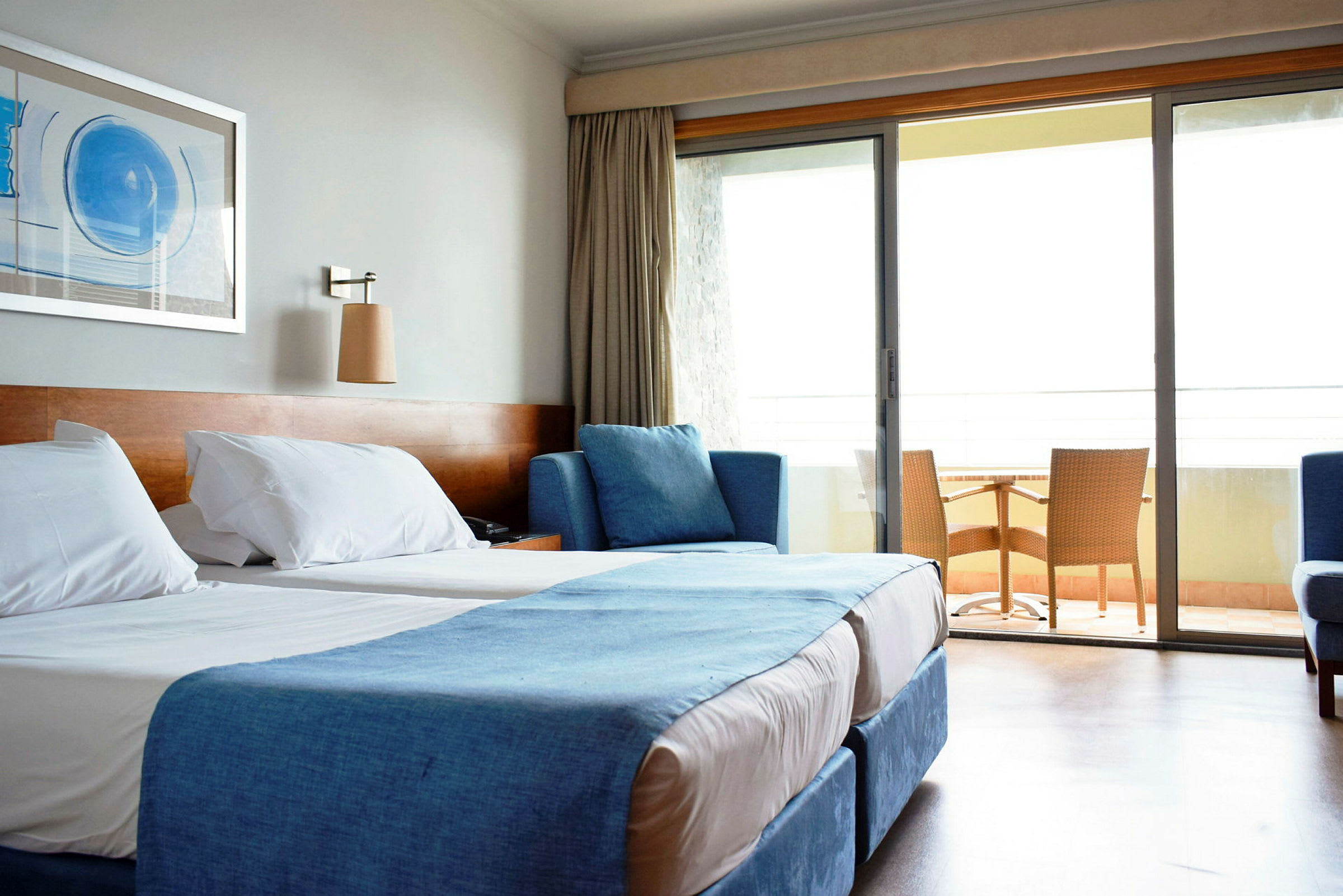 Ett hotellrum med två sängar, blå inredning och en glasad balkongdörr som leder ut till en balkong med bord och stolar. (AI-genererad text)