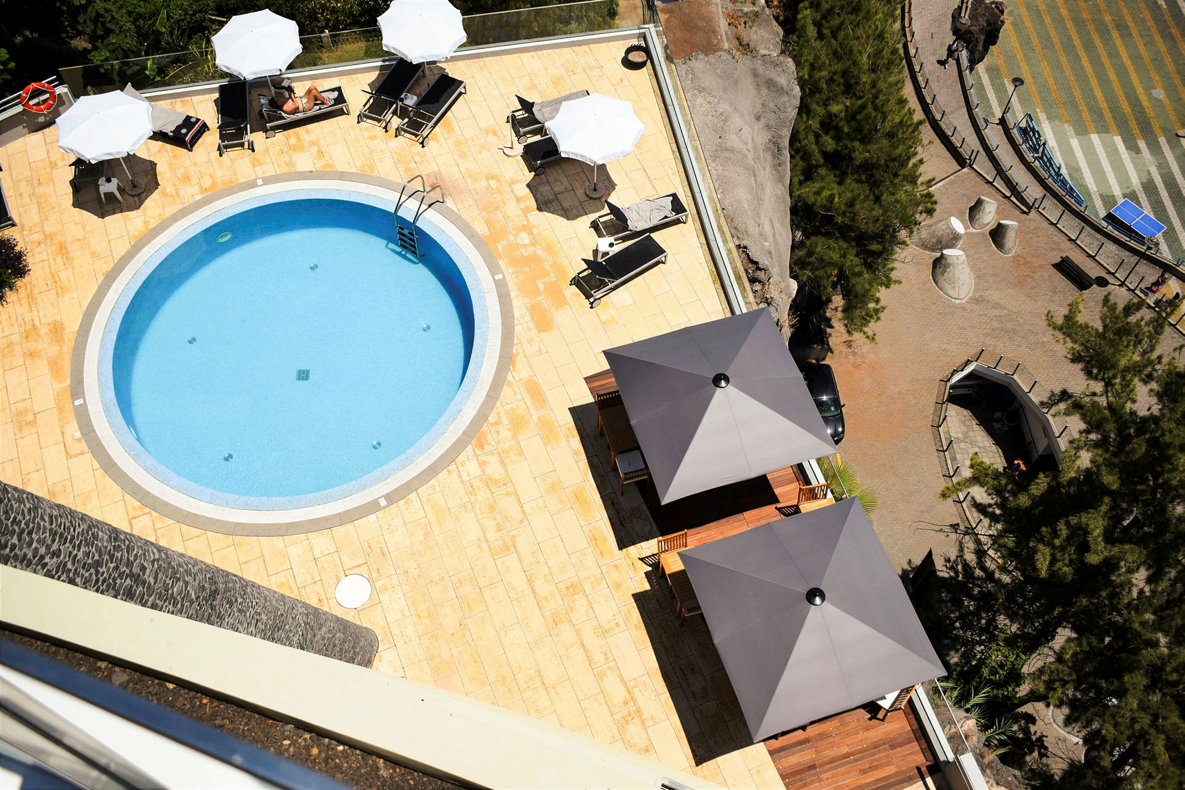 En oval pool omgiven av vit sten som ligger på en solterrass med parasoller och solstolar. (AI-genererad text)