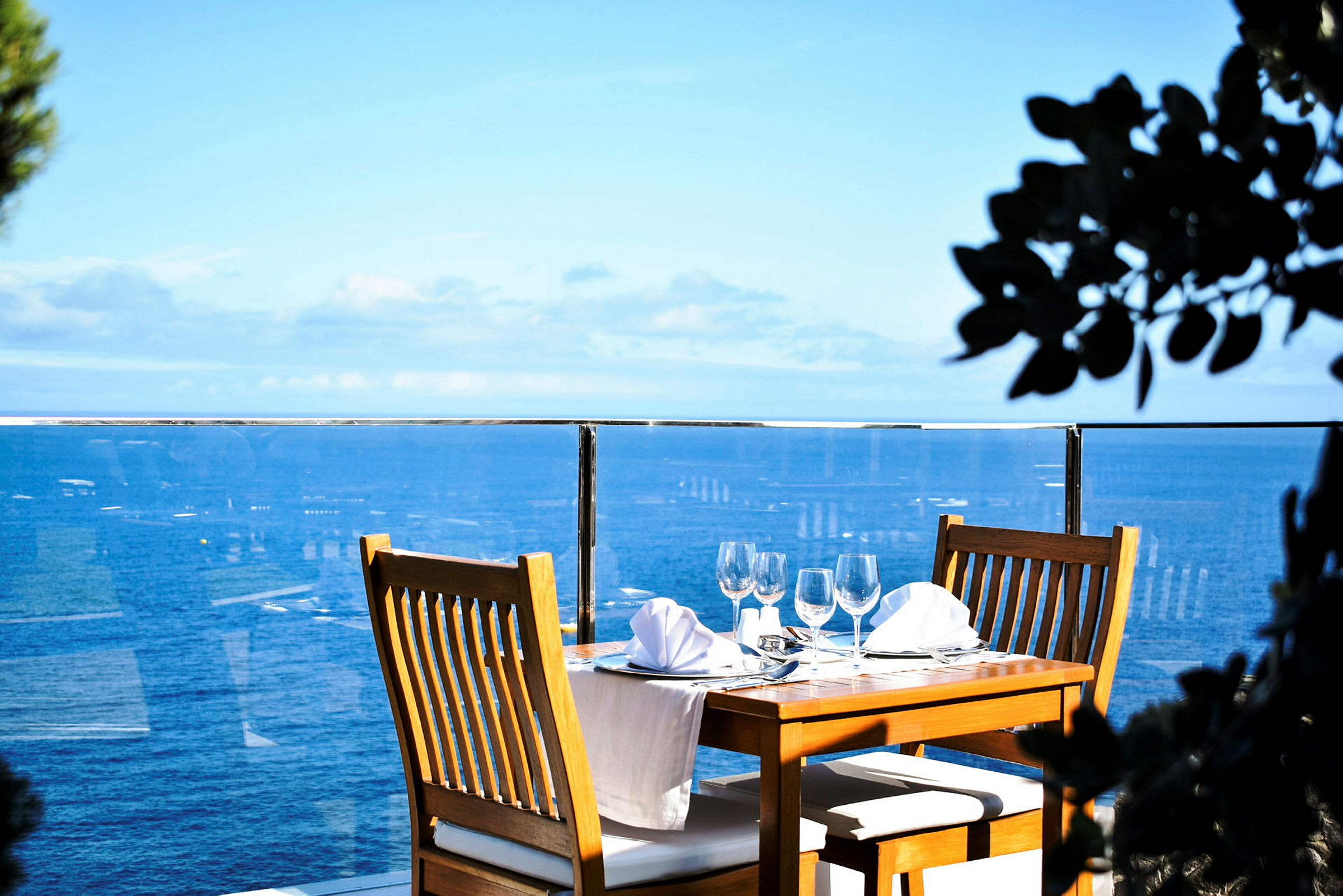 Ett dukat bord med trästolar och vit duk vid en glasräcke med utsikt över havet. (AI-genererad text)