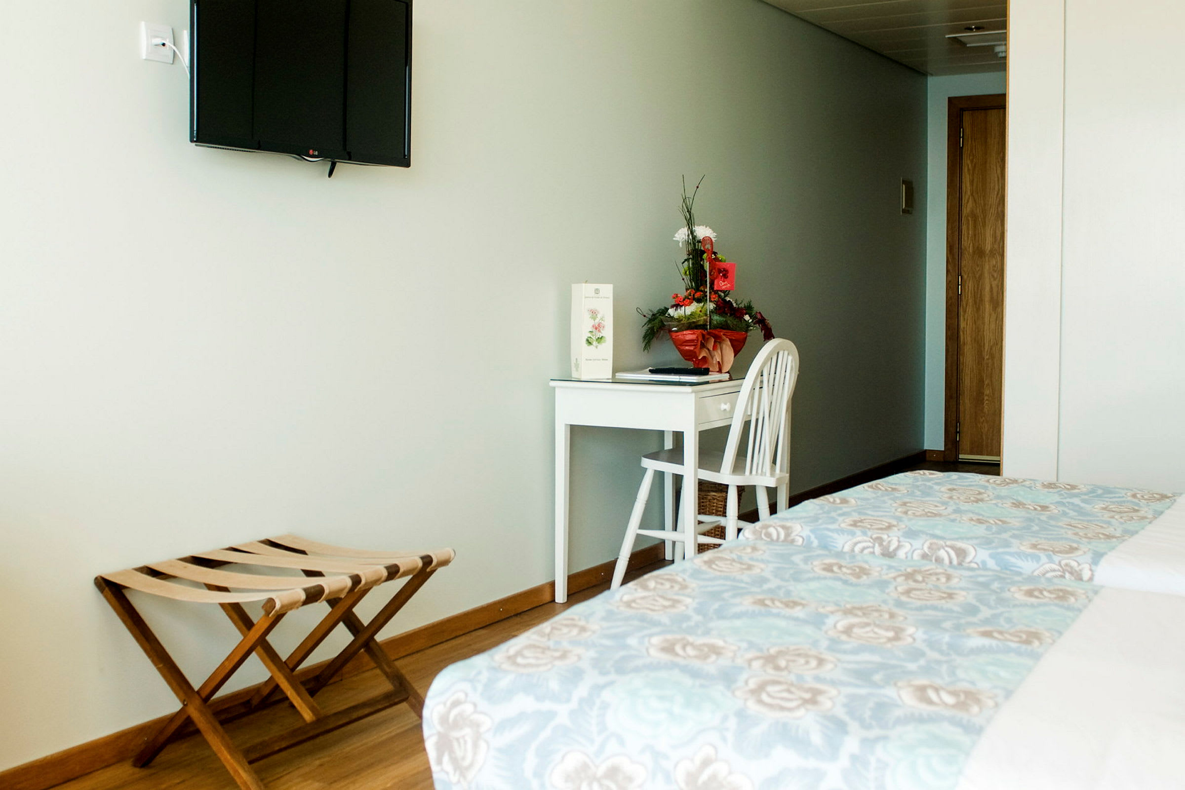 Ett hotellrum med en väggmonterad TV, en vit skrivbord med en stol och en blomsterarrangemang. (AI-genererad text)