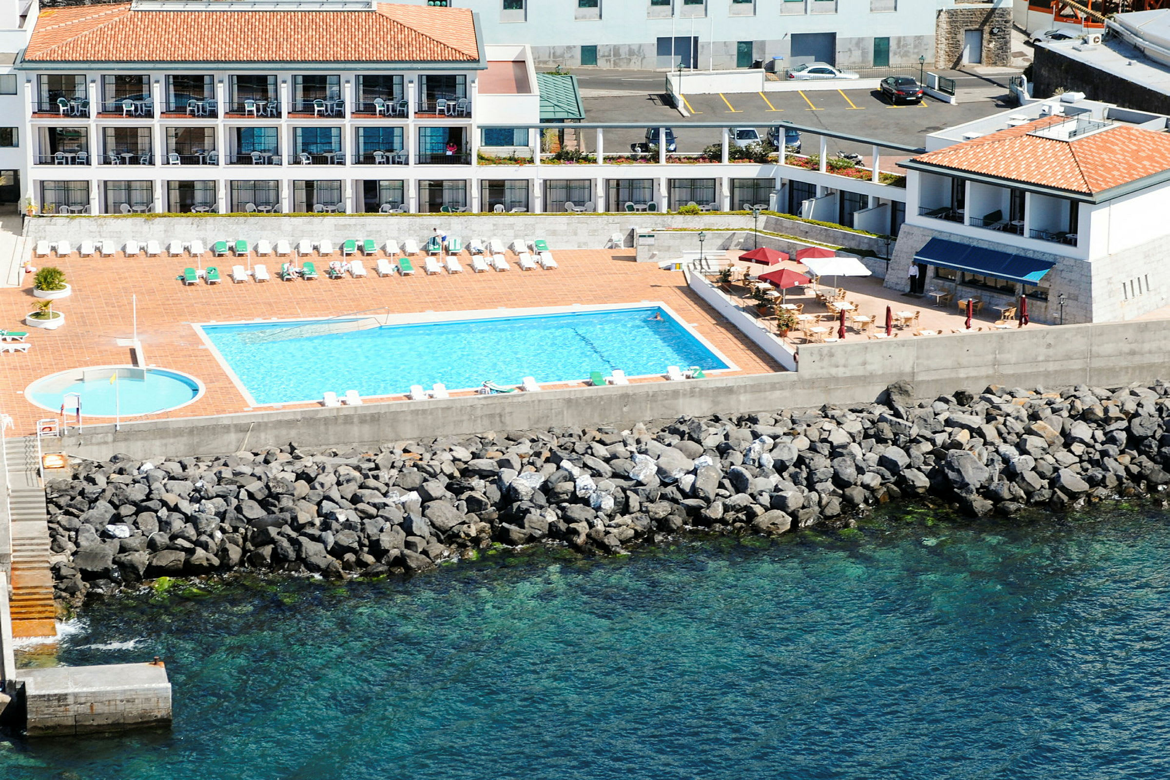 En pool vid havet med solstolar och ett hotell nära. (AI-genererad text)