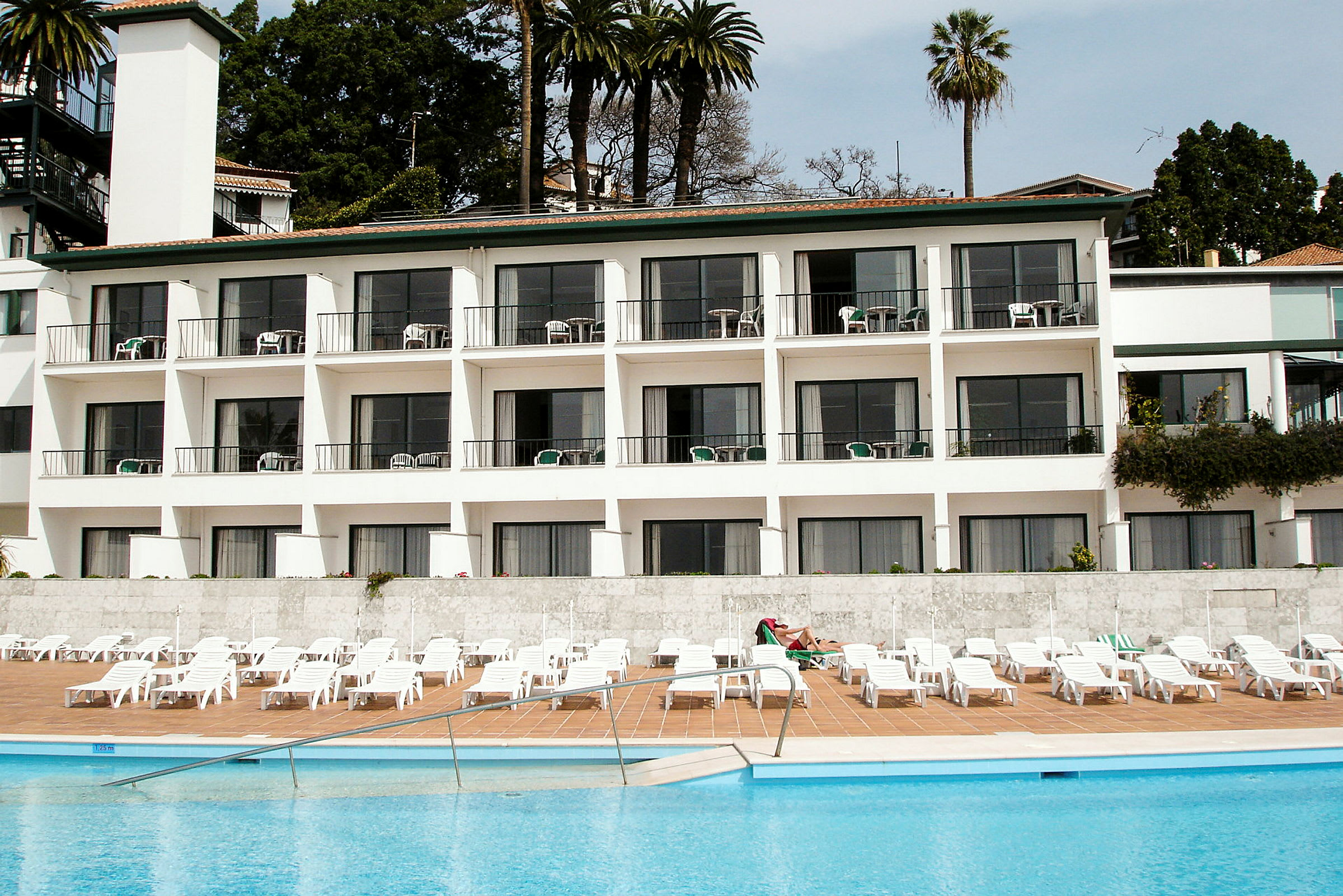 En hotellbyggnad med balkonger framför en pool och solstolar. (AI-genererad text)