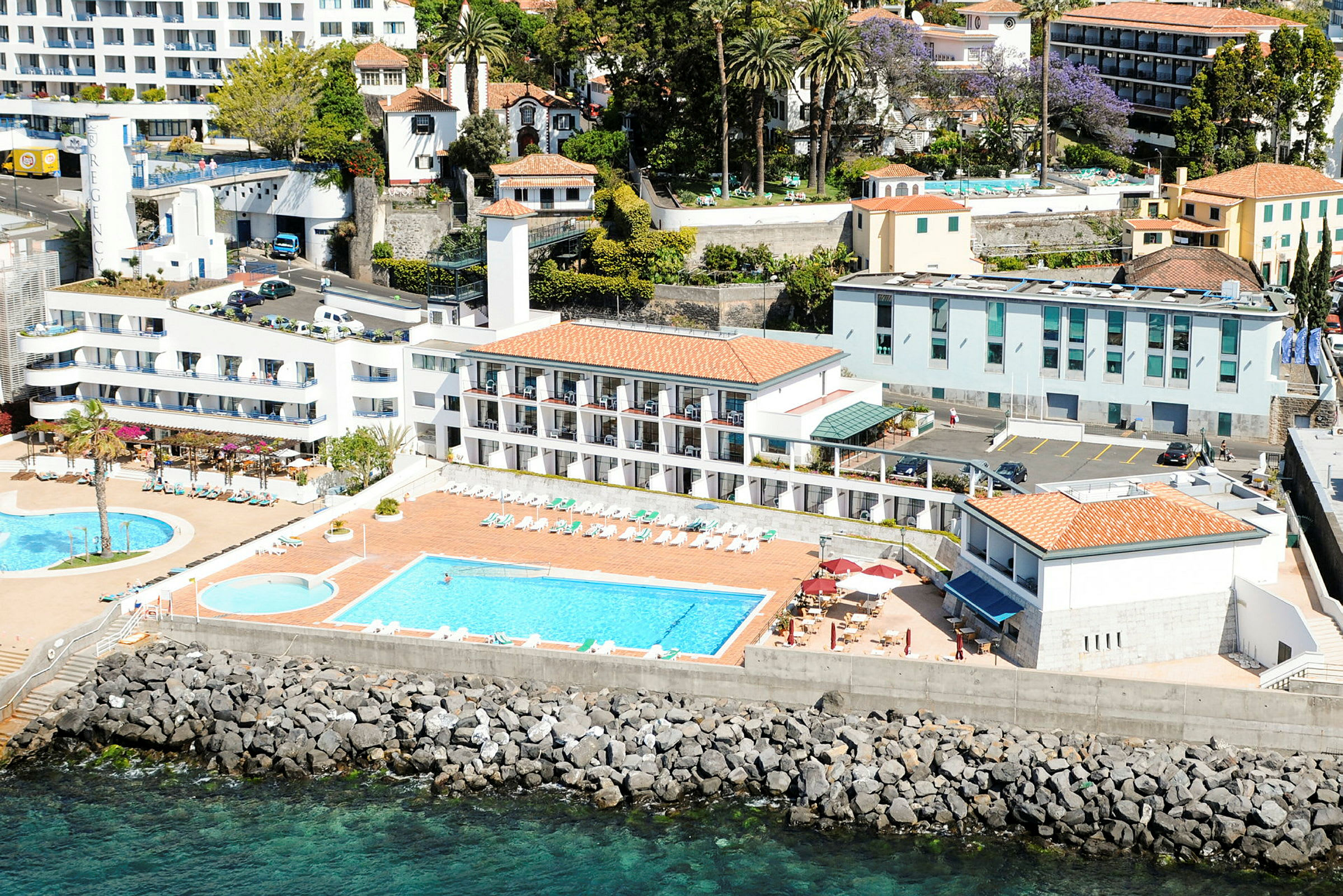 Luftfoto av ett hotell med pooler, solstolar och byggnader nära havet. (AI-genererad text)