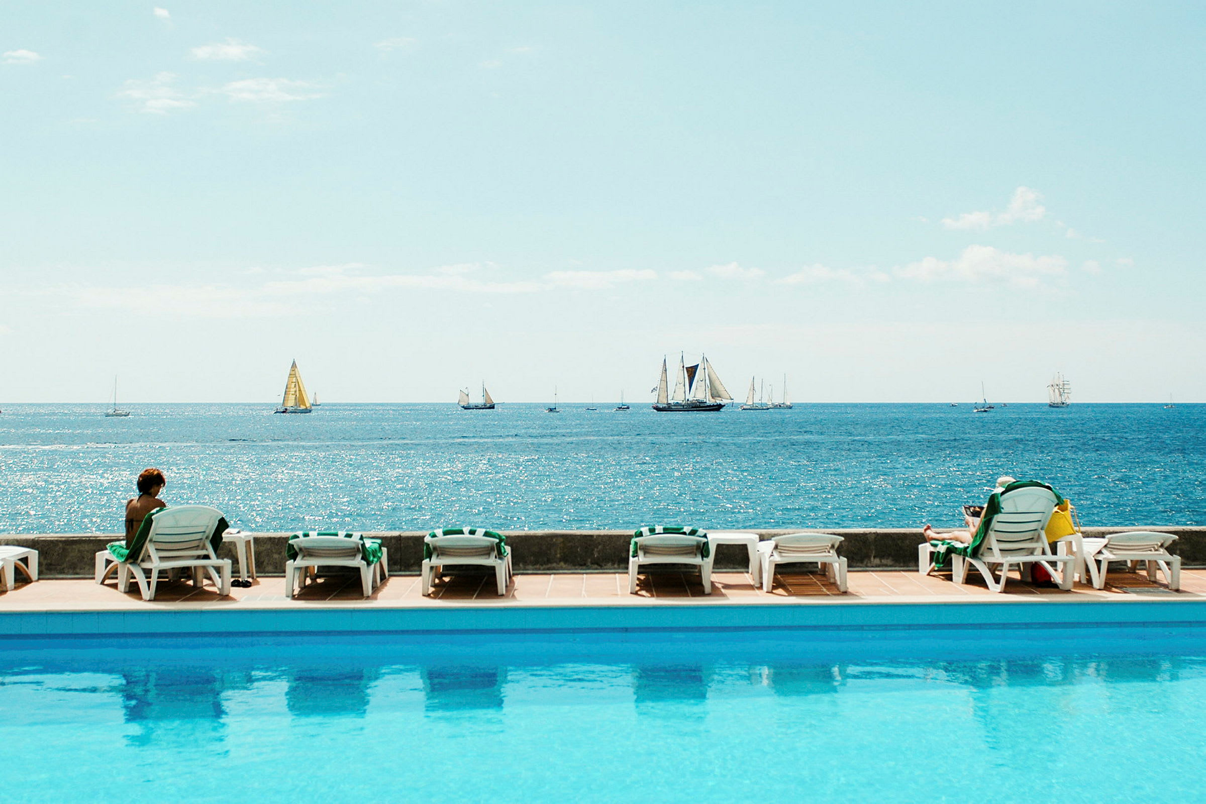 Utsikt över en pool, solstolar och havet med segelbåtar i bakgrunden. (AI-genererad text)
