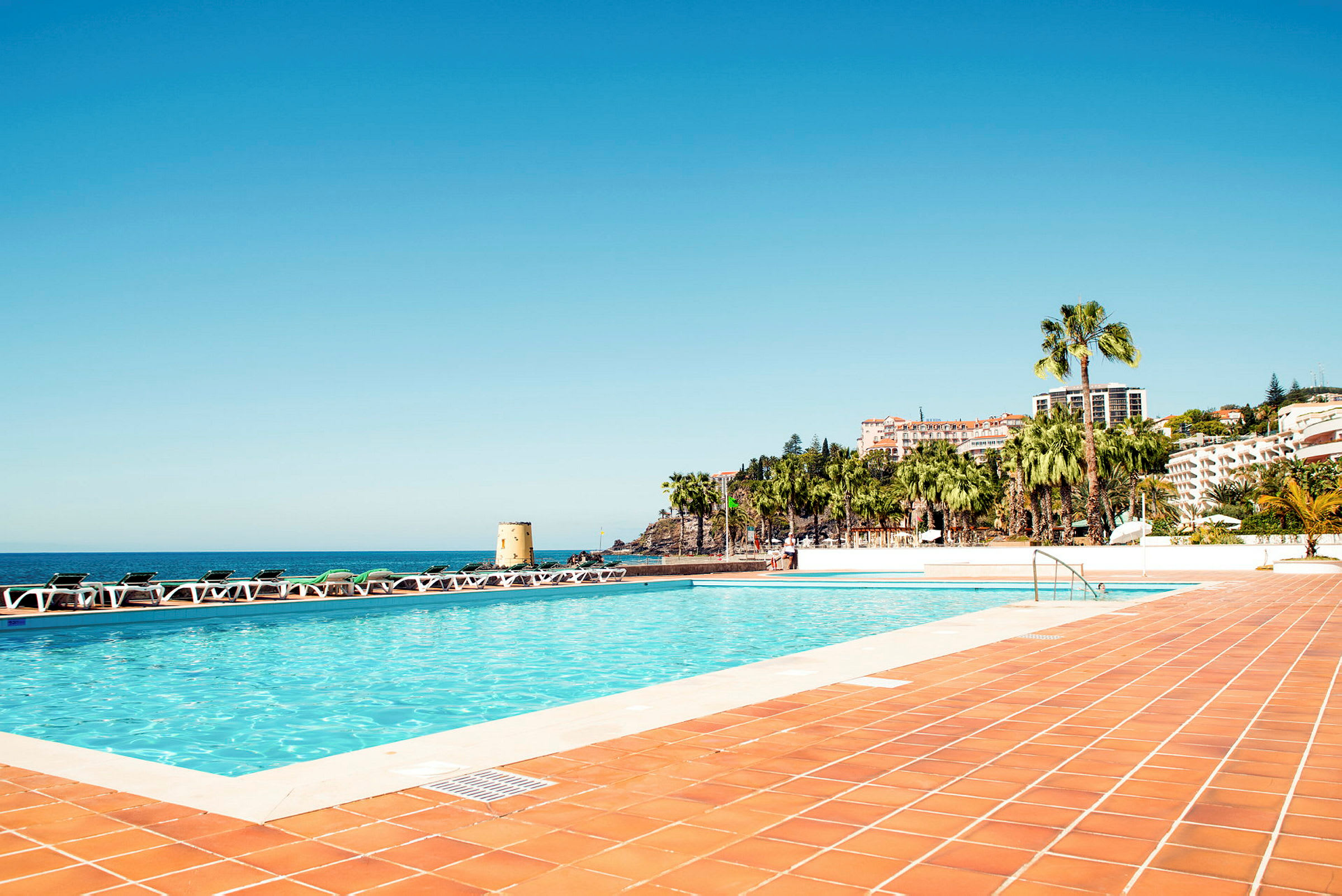 En swimmingpool med omgivande solsängar och vy över havet och bebyggelse med palmer. (AI-genererad text)