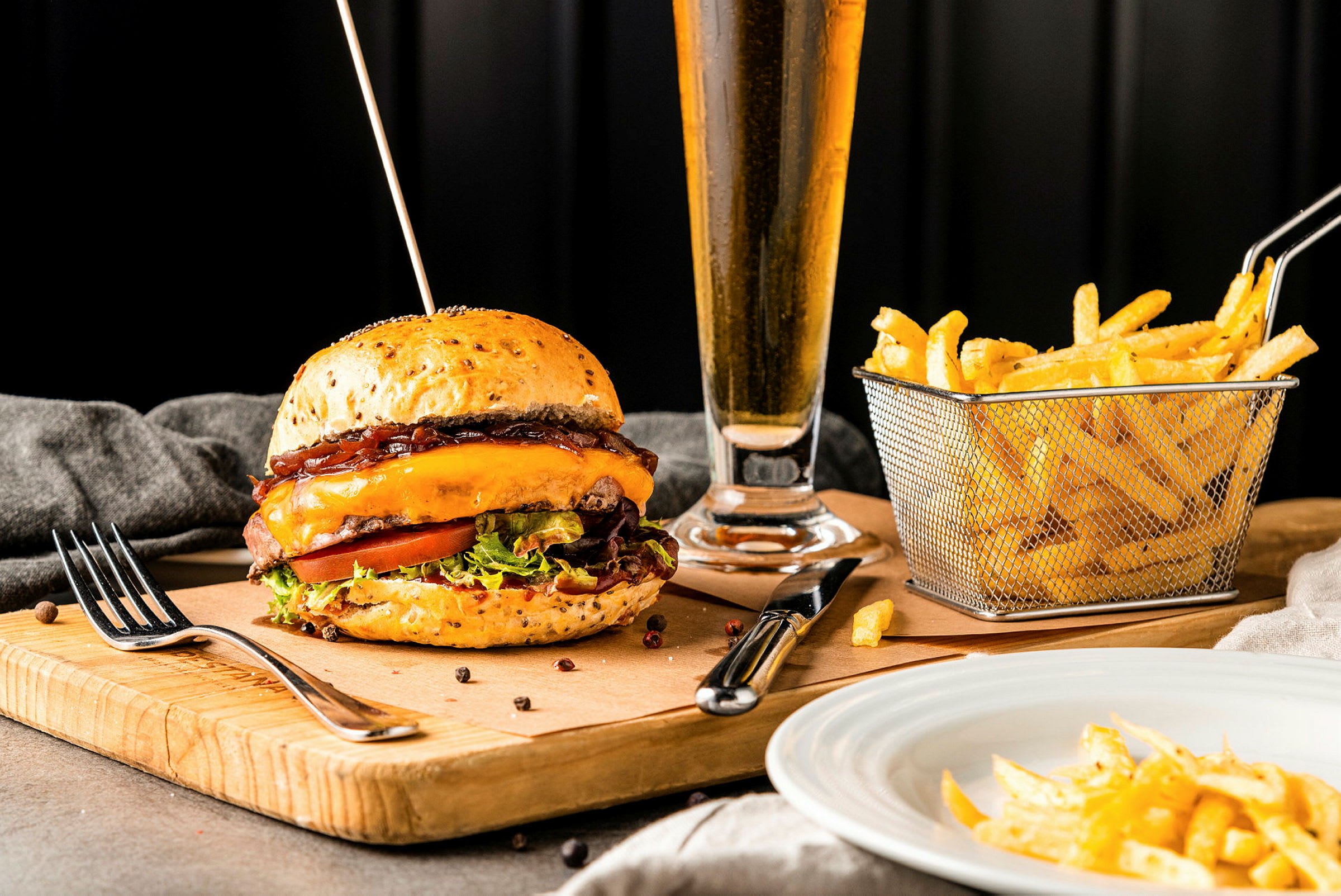 En hamburgare med ost och sallad på en träbräda, med pommes frites i en korg och ett glas dryck i bakgrunden. (AI-genererad text)