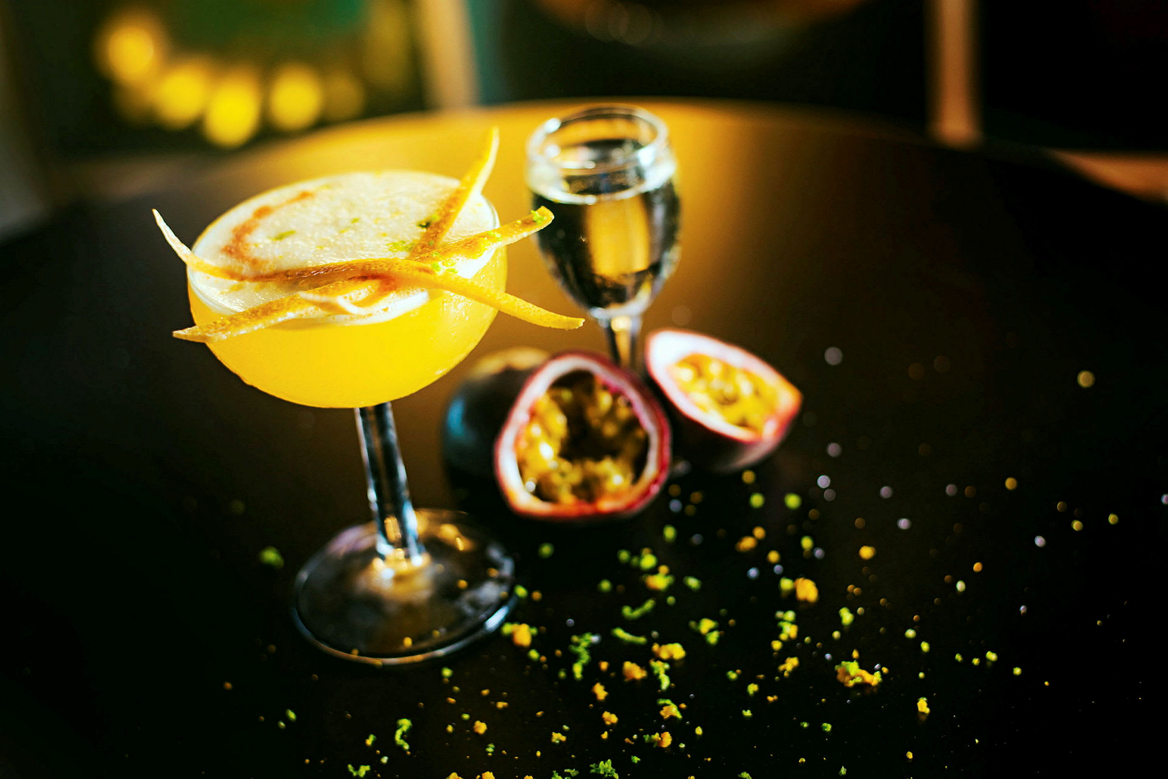 En gul cocktail med dekorationer på ett bord, bredvid ett glas och halverad passionsfrukt. (AI-genererad text)