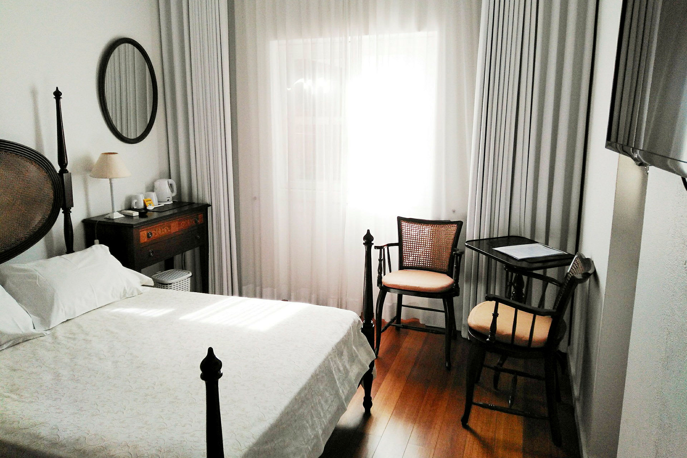 Ett hotellrum med en säng, skrivbord och stol, samt ett fönster med vita gardiner. (AI-genererad text)
