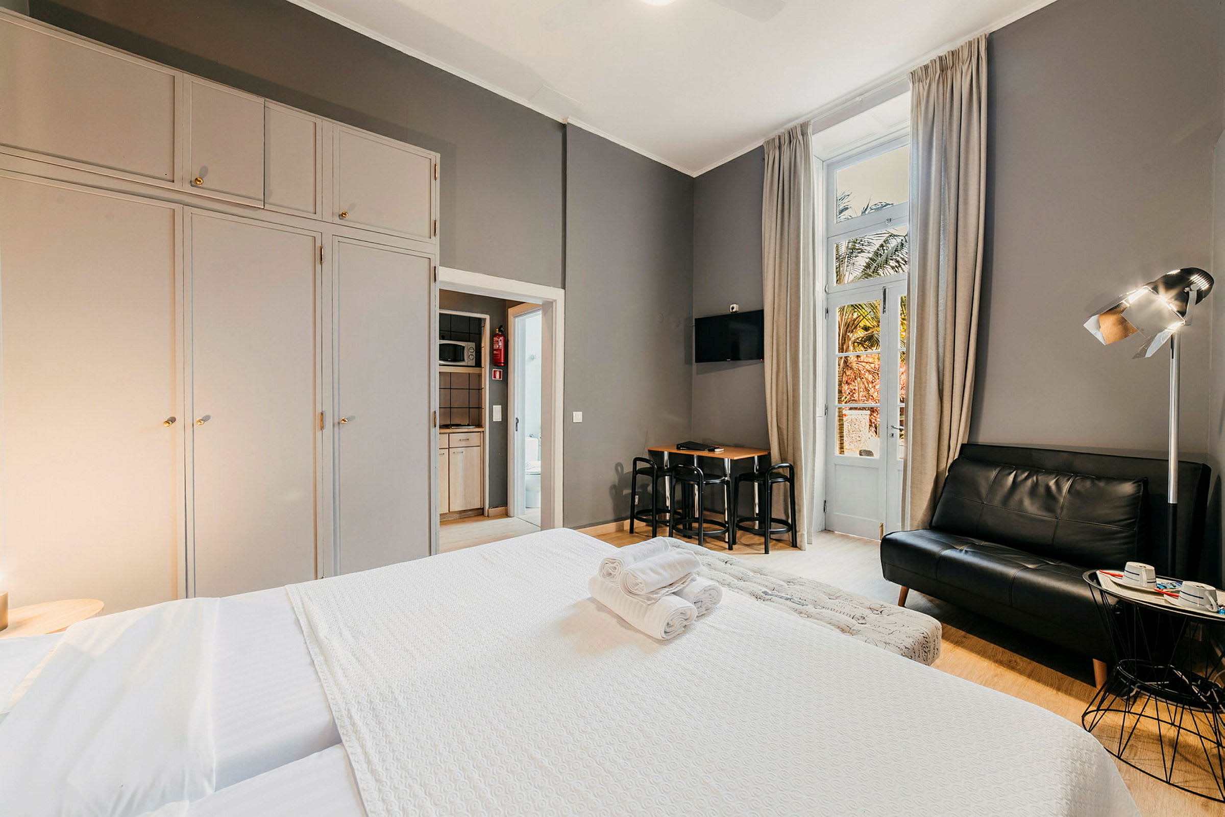 Hotellrum med säng, soffa och matbord, stora garderober samt en balkongdörr. (AI-genererad text)