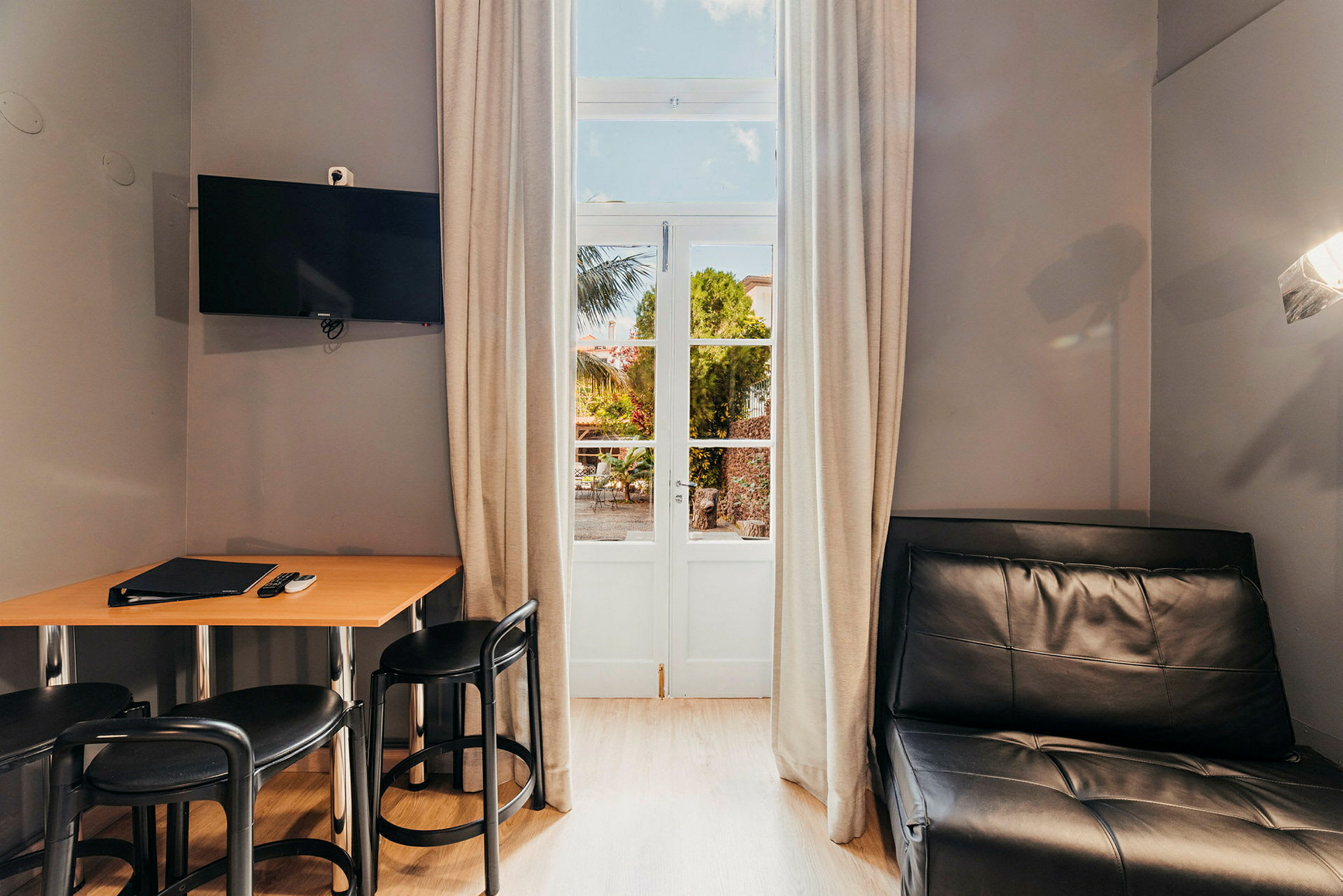 Ett rum med ett bord, stolar, en svart soffa och en TV på väggen, samt en dörr med glaspartier och gardiner. (AI-genererad text)