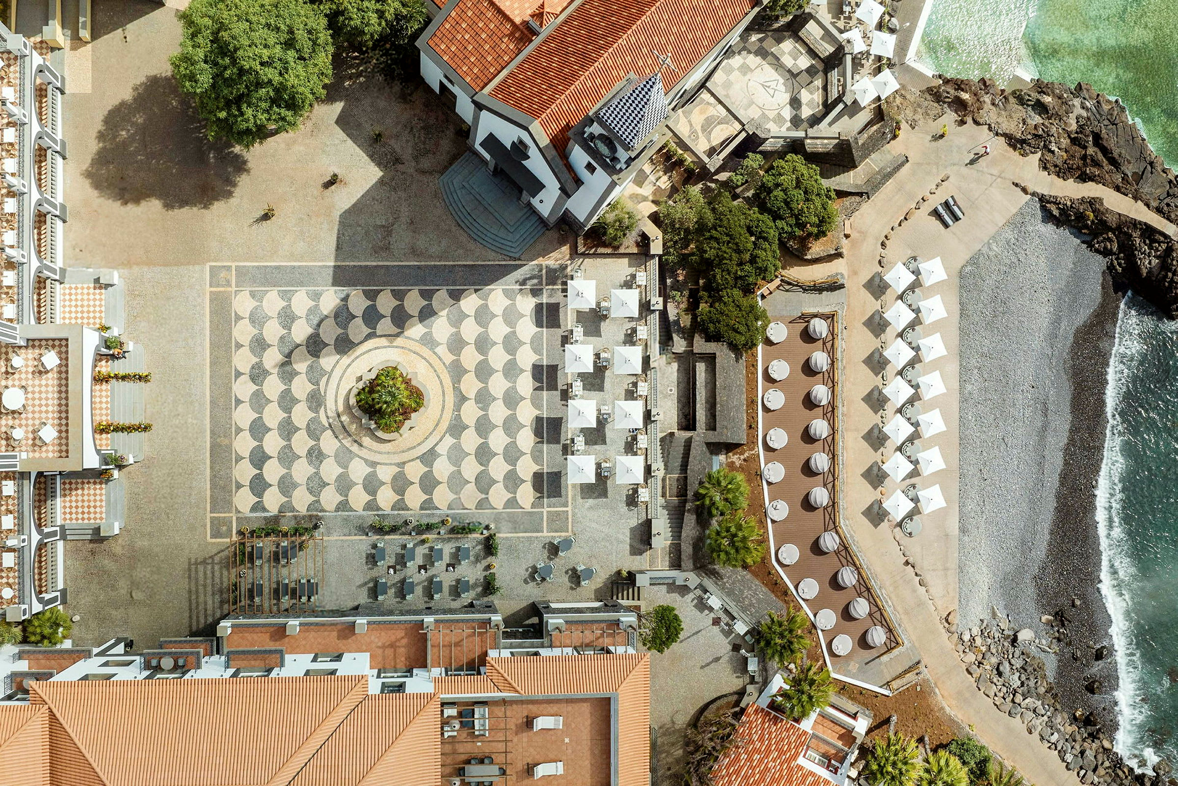 Flygfoto över en torg med mönstrat stenlagt golv, omgärdat av byggnader med tegeltak och en strandlinje. (AI-genererad text)