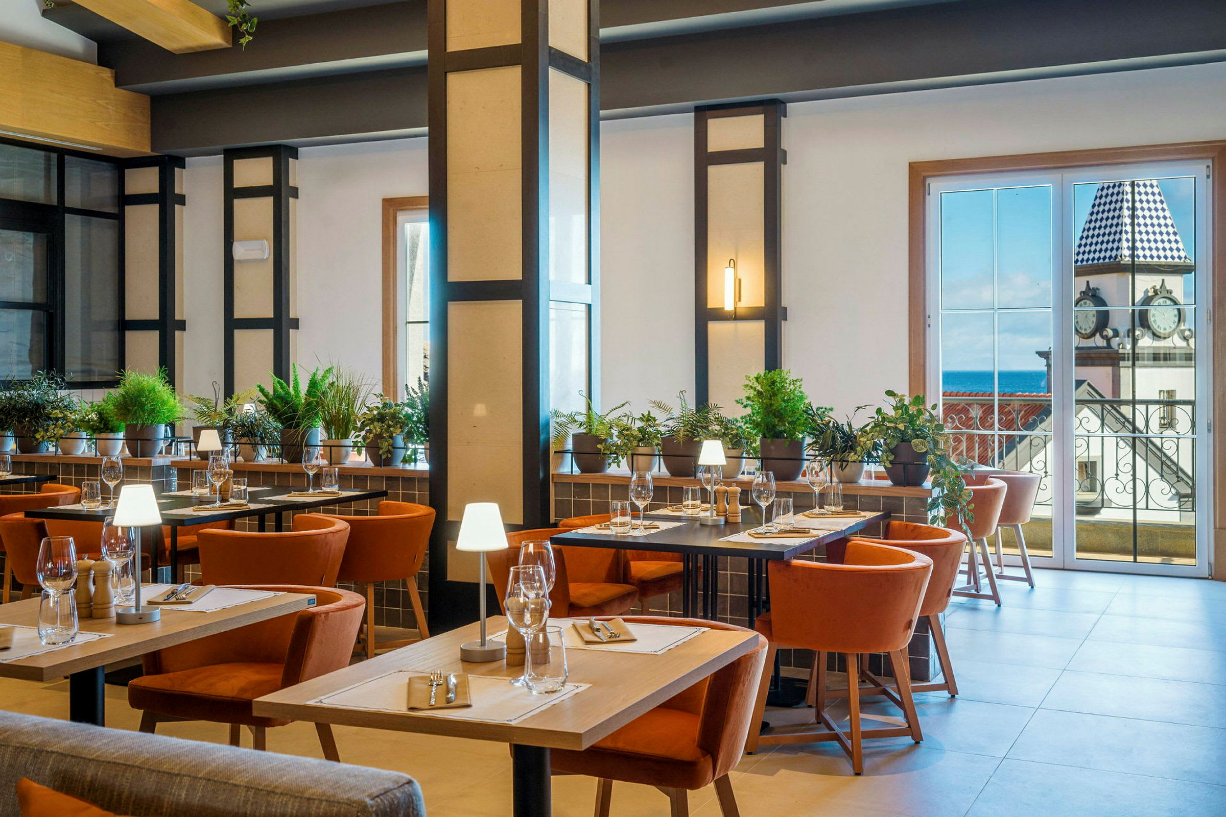 Ett välbelyst restauranginteriör med bord, stolar och grön växtdekoration. I bakgrunden finns stora fönster som visar en vy av en byggnad med ett klocktorn. (AI-genererad text)