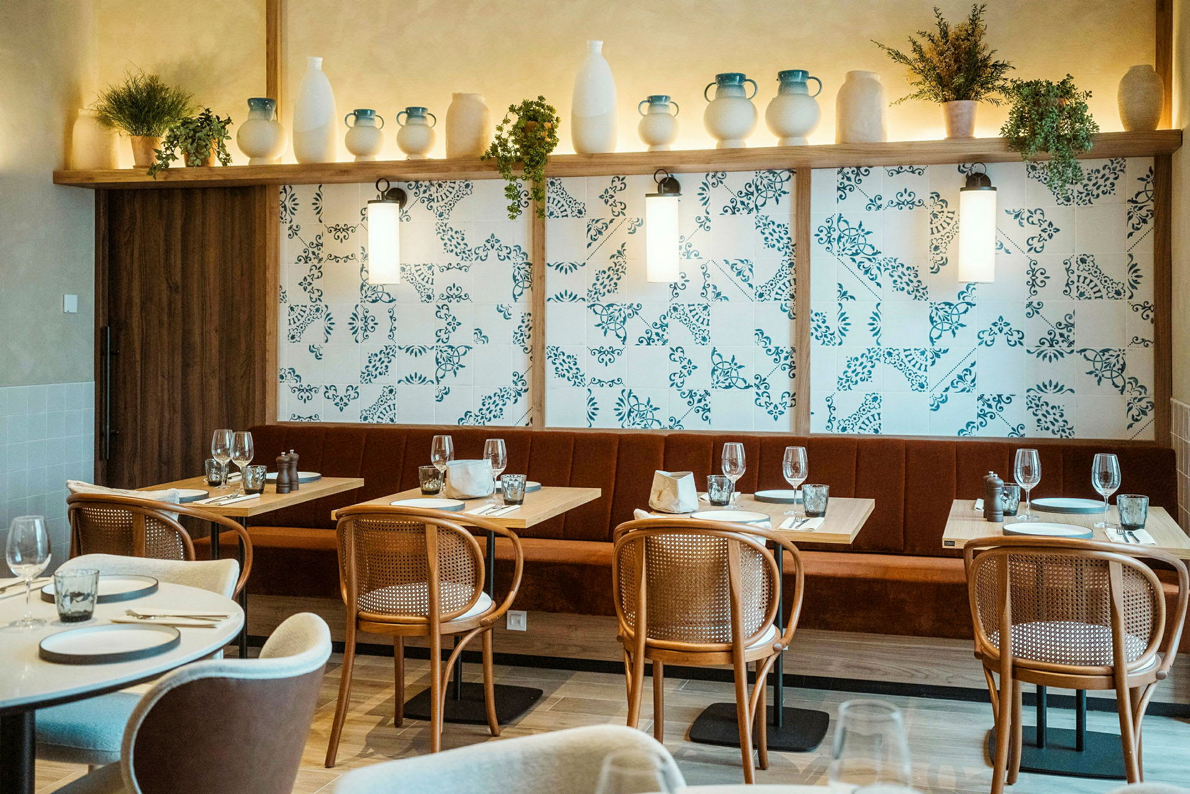 En interiörbild av en restaurang med dukade bord, stolar och en dekorerad vägg med hyllor fyllda med keramik och växter. (AI-genererad text)