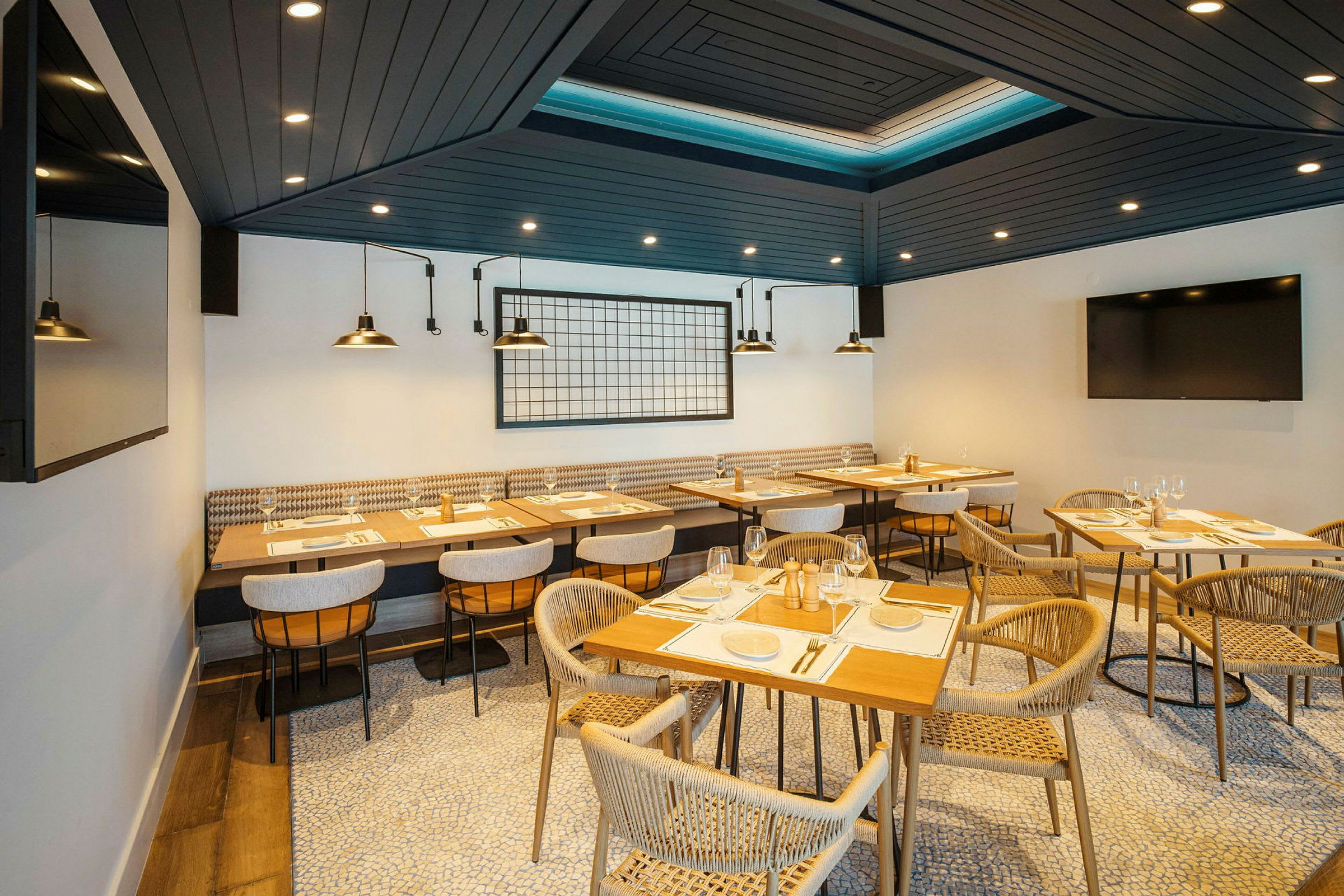 En modern restaurang med träbord och bekväma stolar arrangerade på ett organiskt mönstrat golv. Taket är mörkt med spots, och bakre väggen har industriella lampor och dekor. (AI-genererad text)