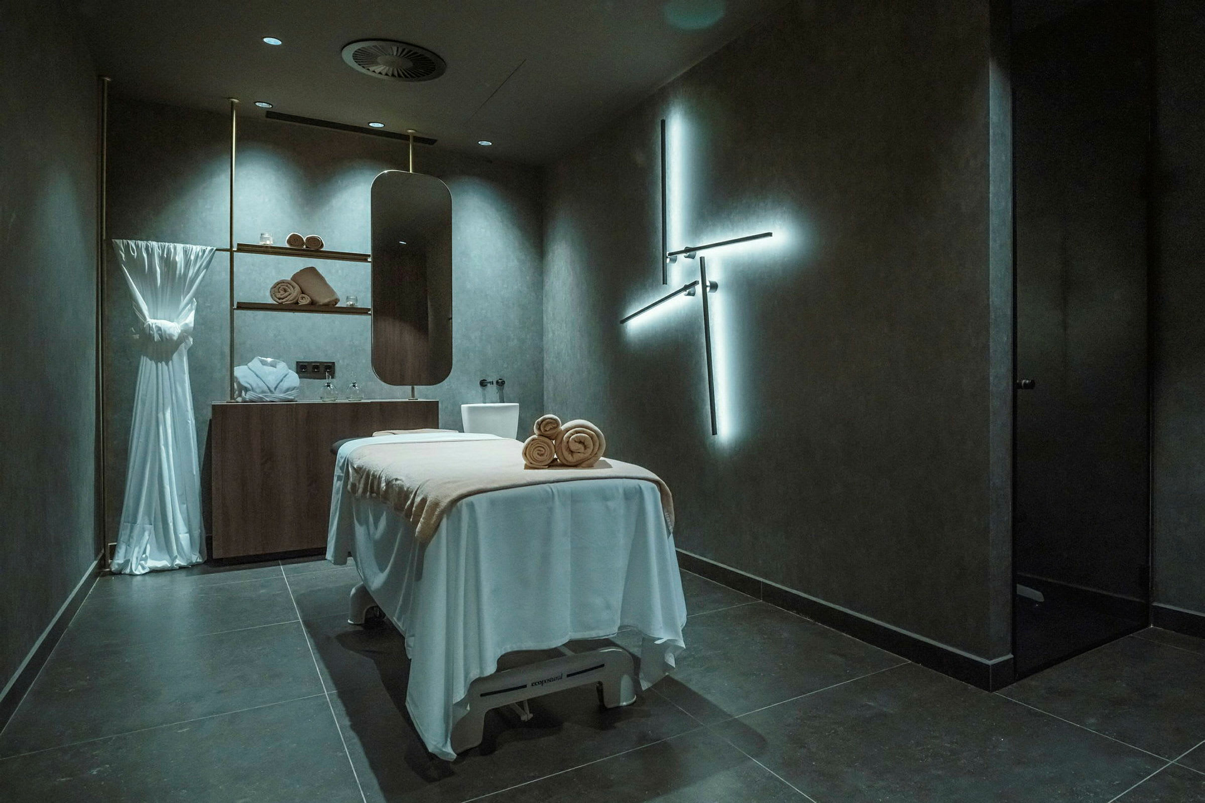 Ett rum med en massagebänk klädd i vitt, hyllor med handdukar och dekoration samt en spegel på väggen. Rummet har en modern och avkopplande atmosfär. (AI-genererad text)