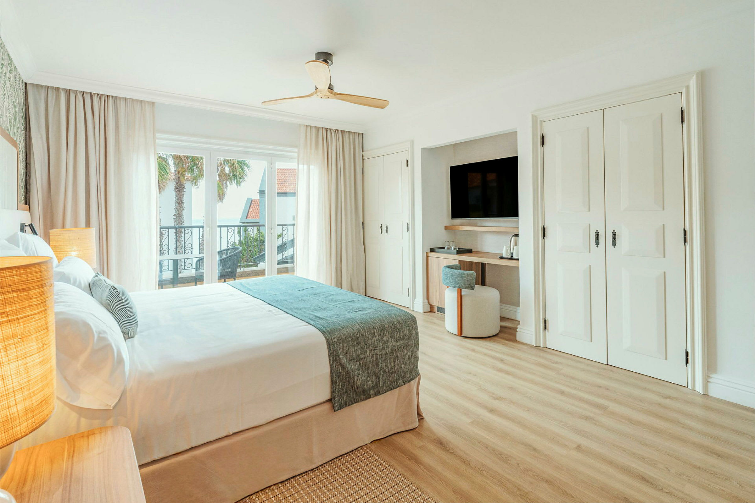 Ett ljus, modernt hotellrum med en dubbelsäng, stor fönstervy, balkong och ett välordnat skrivbord med TV. (AI-genererad text)