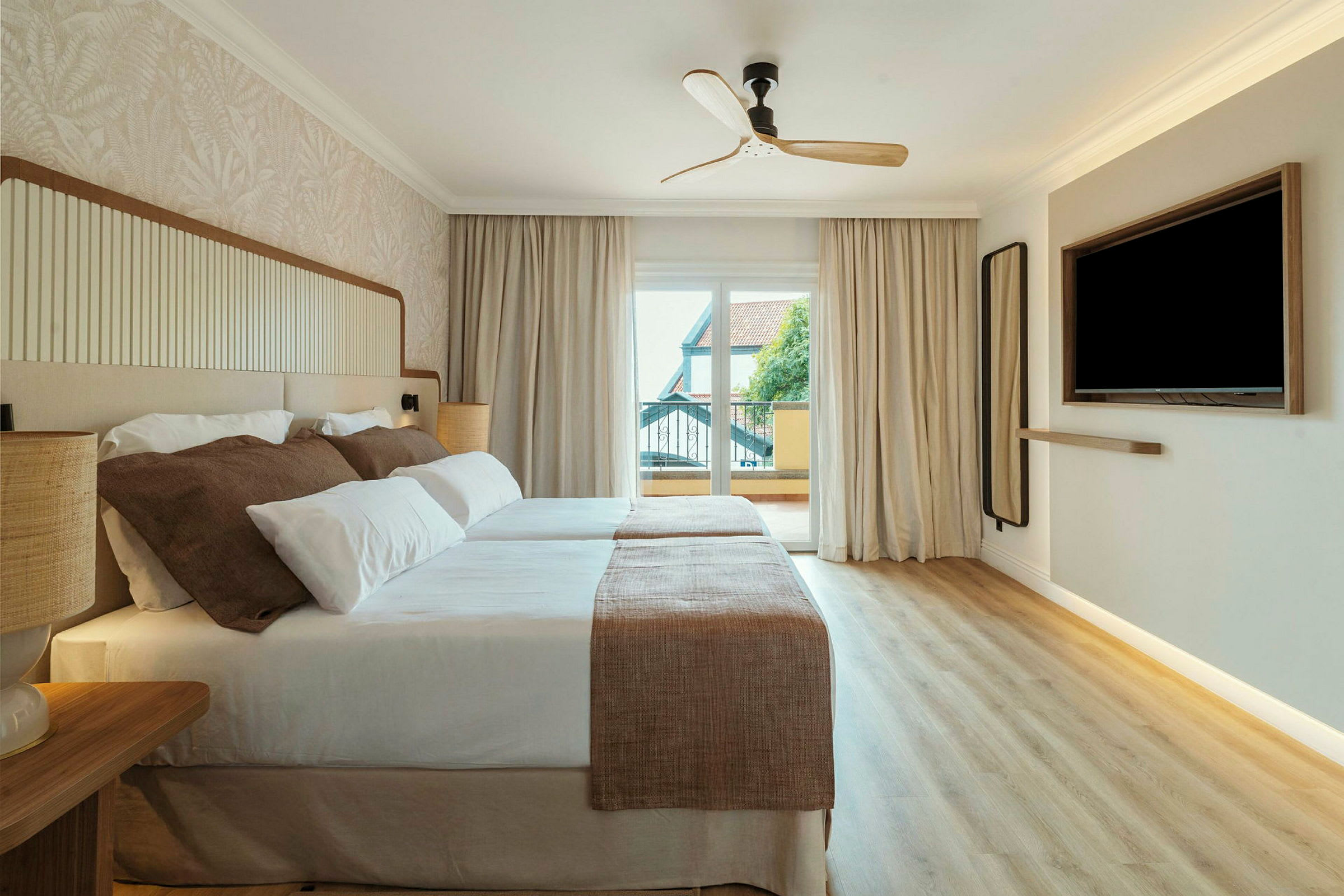 Ett hotellrum med två sängar som täcks av vita och beige sängkläder. Det finns en takfläkt, ett stort fönster med utsikt och en väggmonterad TV. (AI-genererad text)