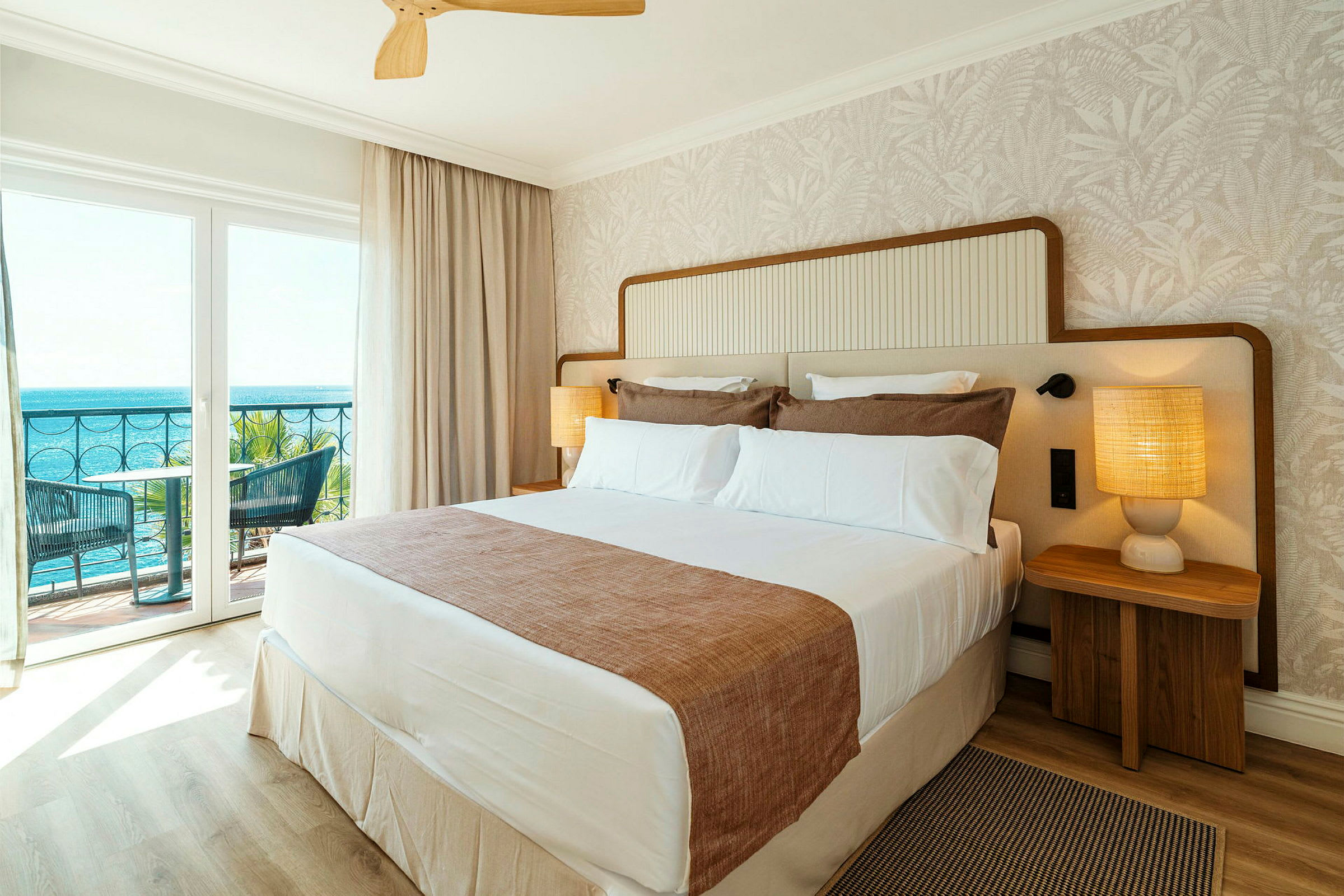 Ett hotellrum med en stor säng, två sängbord med lampor och en balkong med utsikt mot havet. (AI-genererad text)