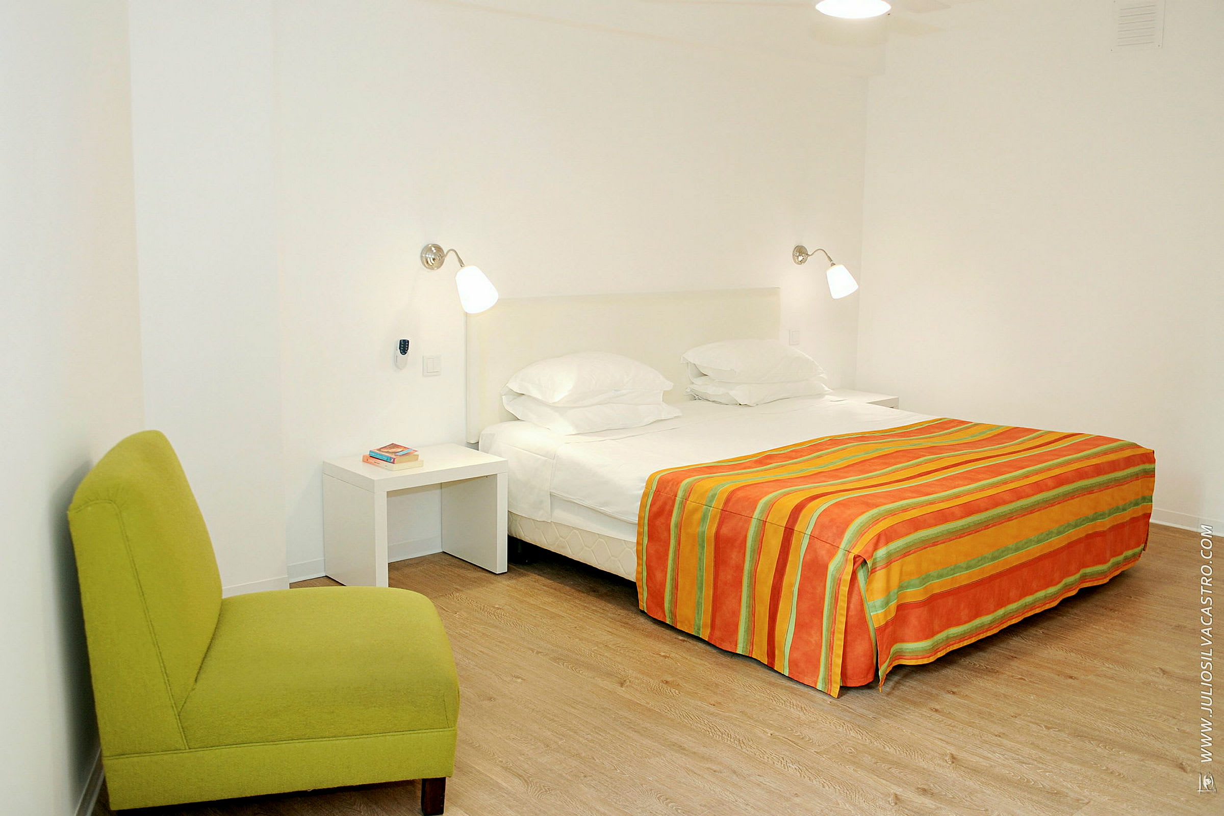 Ett sovrum med en dubbelsäng klädd i en randig orange och grön filt, två sänglampor, ett vitt nattduksbord och en grön stol. (AI-genererad text)