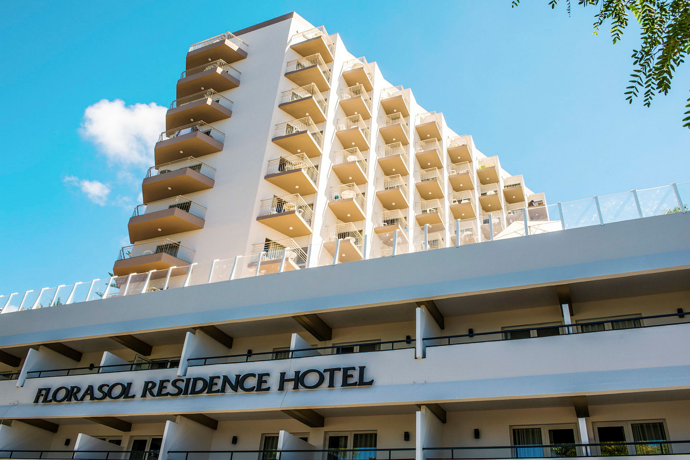 Vy av byggnaden Florasol Residence Hotel med flera balkonger och en blå himmel i bakgrunden. (AI-genererad text)