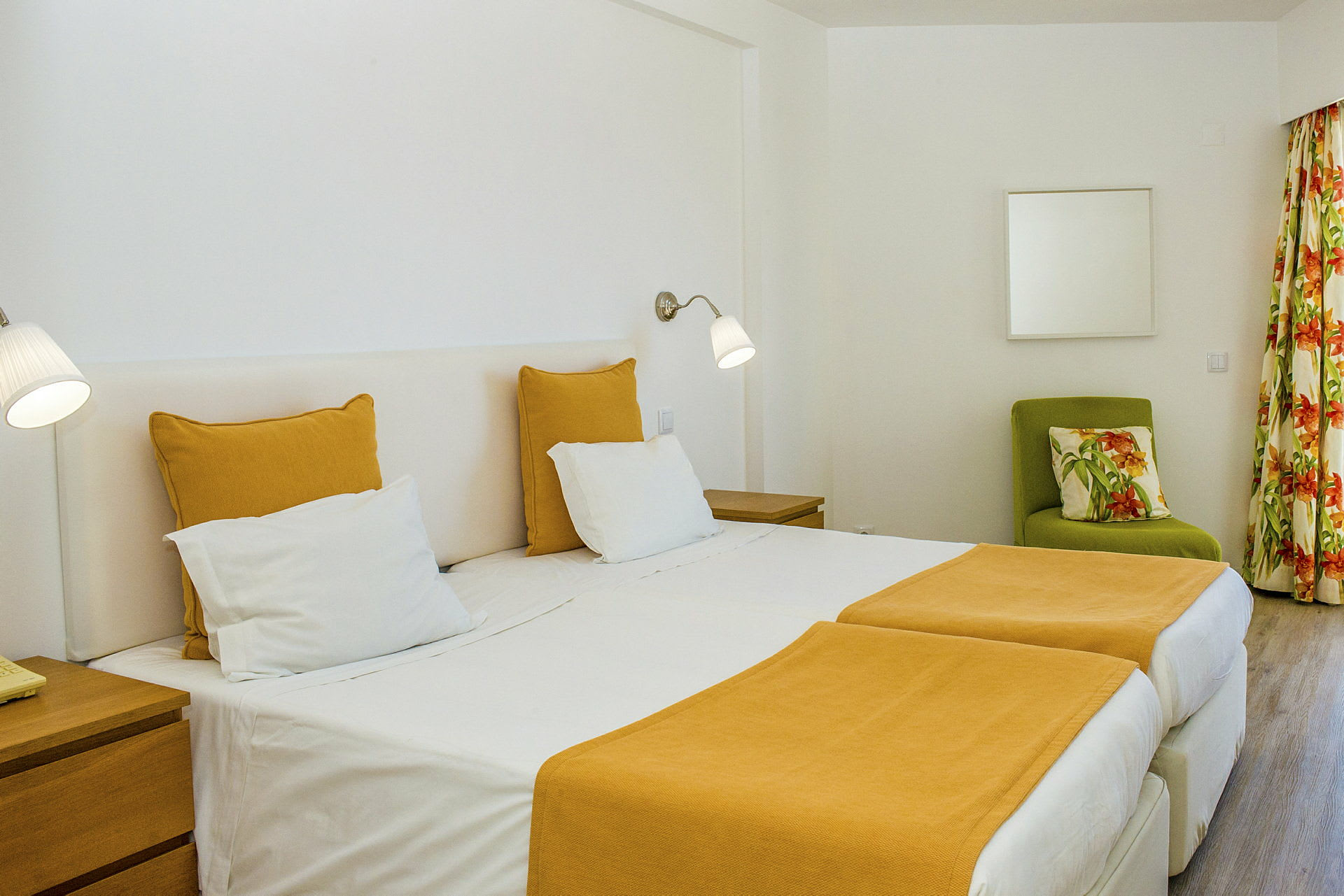 Ett hotellrum med två enkelsängar och gul sängklädsel. Ett grönt fåtölj med blommigt kudde och gardiner syns i bakgrunden. (AI-genererad text)