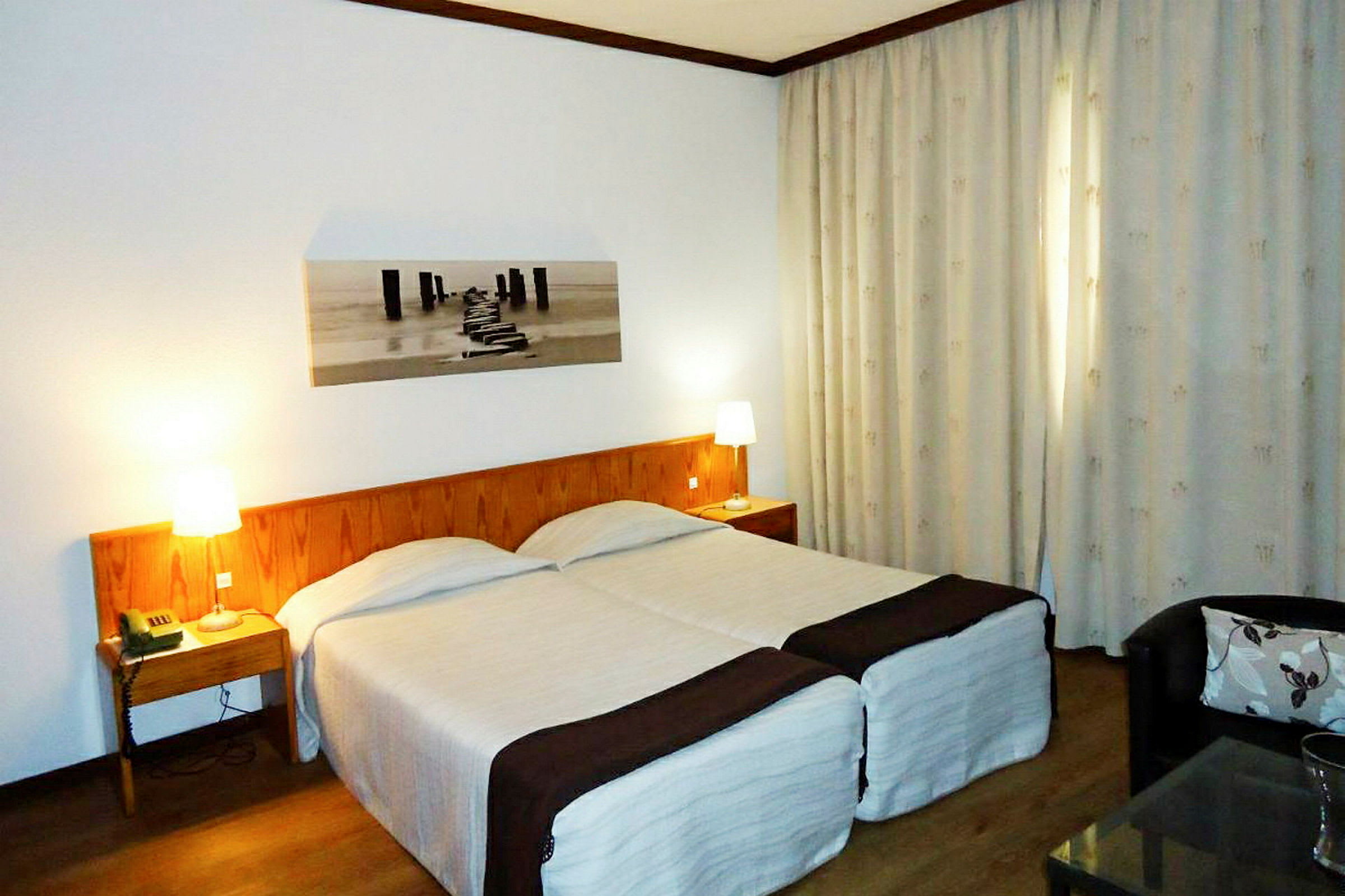 Ett hotellrum med två enkelsängar, ett trähuvudgavel och nattduksbord med lampor. En tavla hänger på väggen och gardiner täcker fönstret. (AI-genererad text)