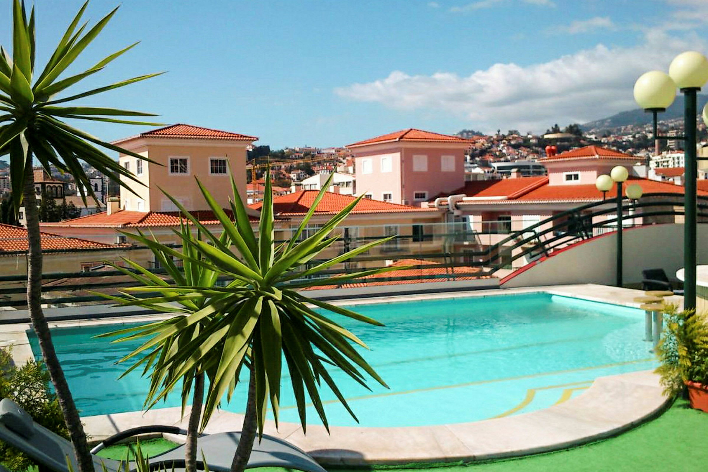 En pool omgiven av grönska och en bakgrund med hus med röda tegeltak. (AI-genererad text)