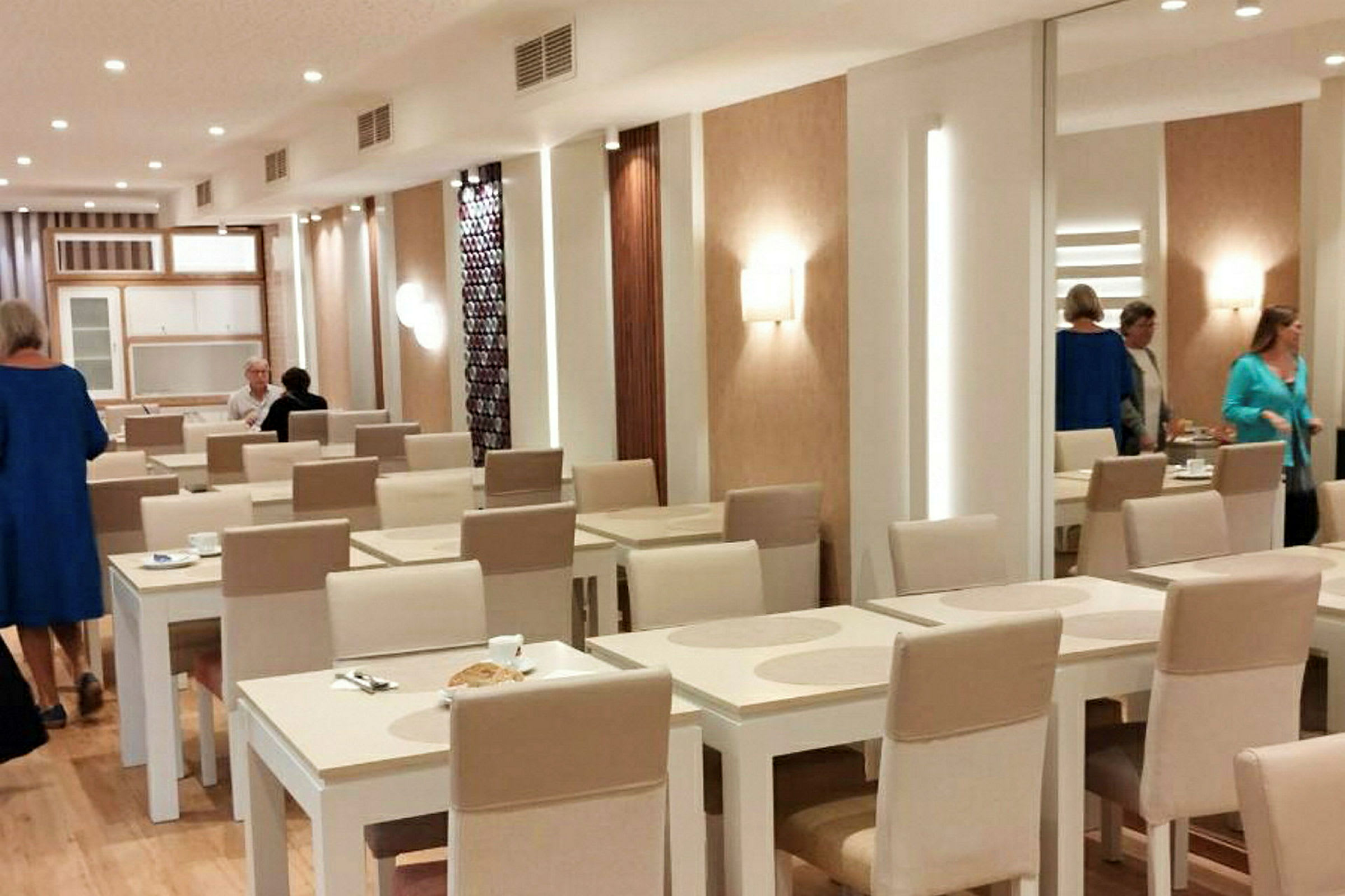 En matsal med vita bord och beige stolar, samt personer som rör sig i bakgrunden. (AI-genererad text)