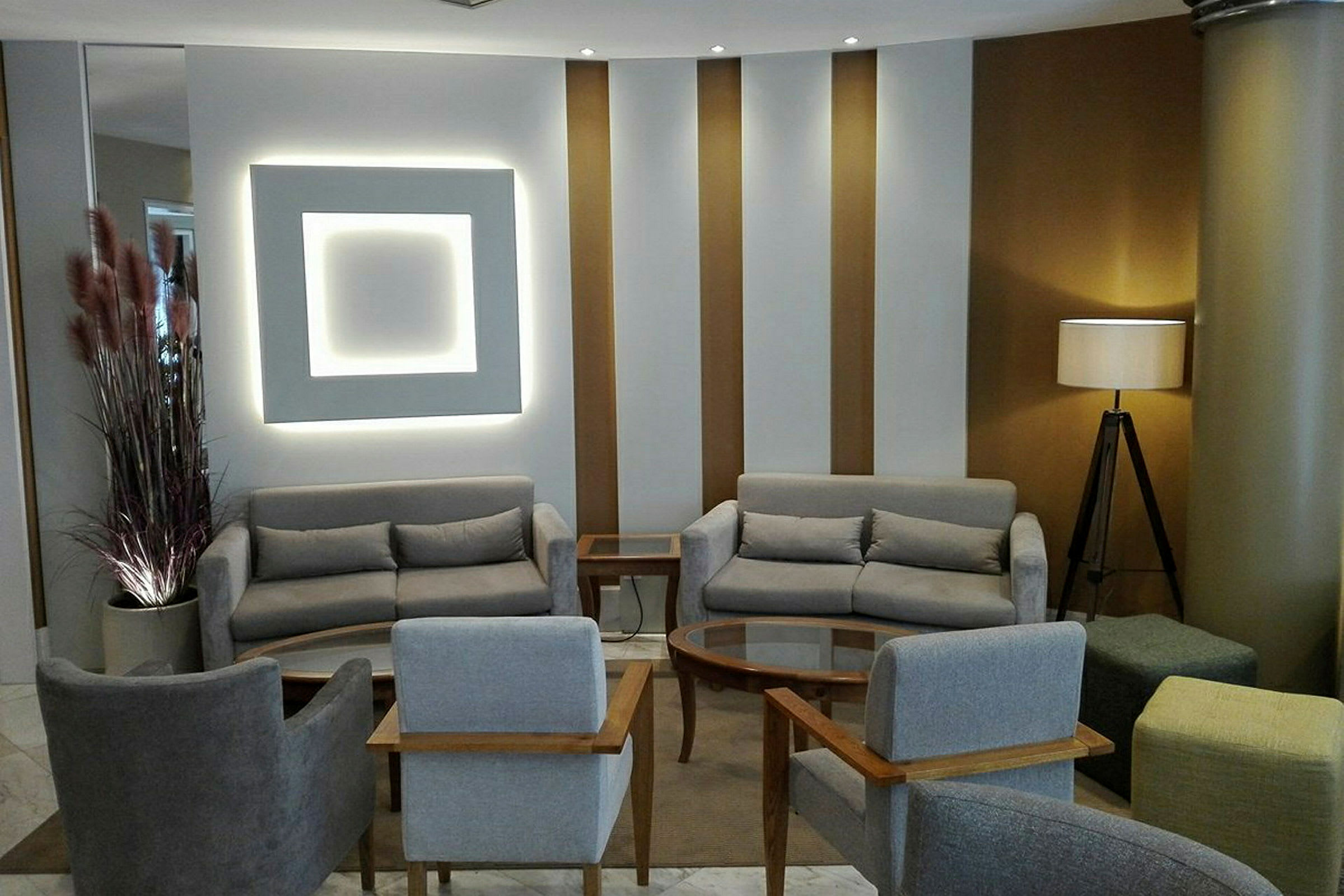 Ett modernt loungeområde med grå soffor och stolar arrangerade kring ett soffbord på en matta. En lampa står i hörnet bredvid en kolonn. (AI-genererad text)