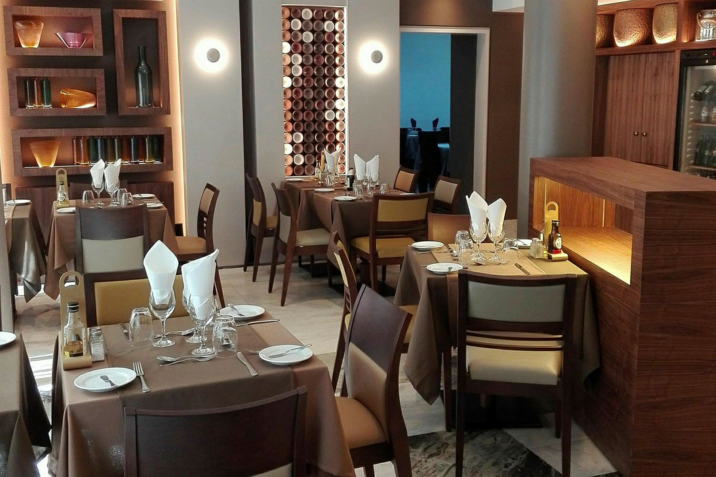 En modern restaurang med bord klädda i bruna dukar och tillhörande trästolar. I bakgrunden finns vägghyllor med dekorativa föremål. (AI-genererad text)