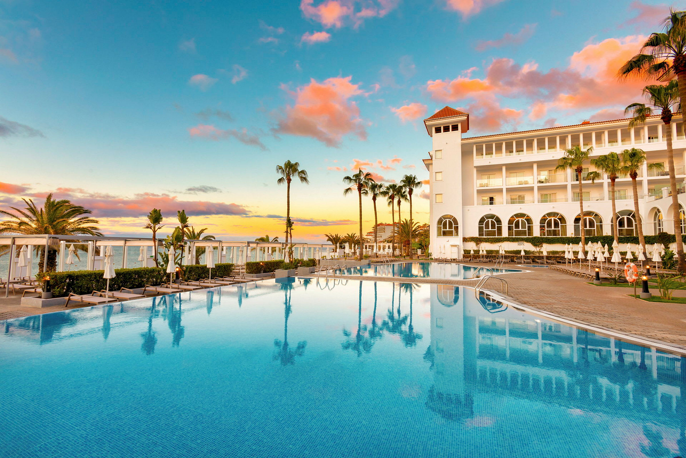 Hotell med stora fönster och pool, omgiven av solstolar och palmer vid solnedgången. (AI-genererad text)