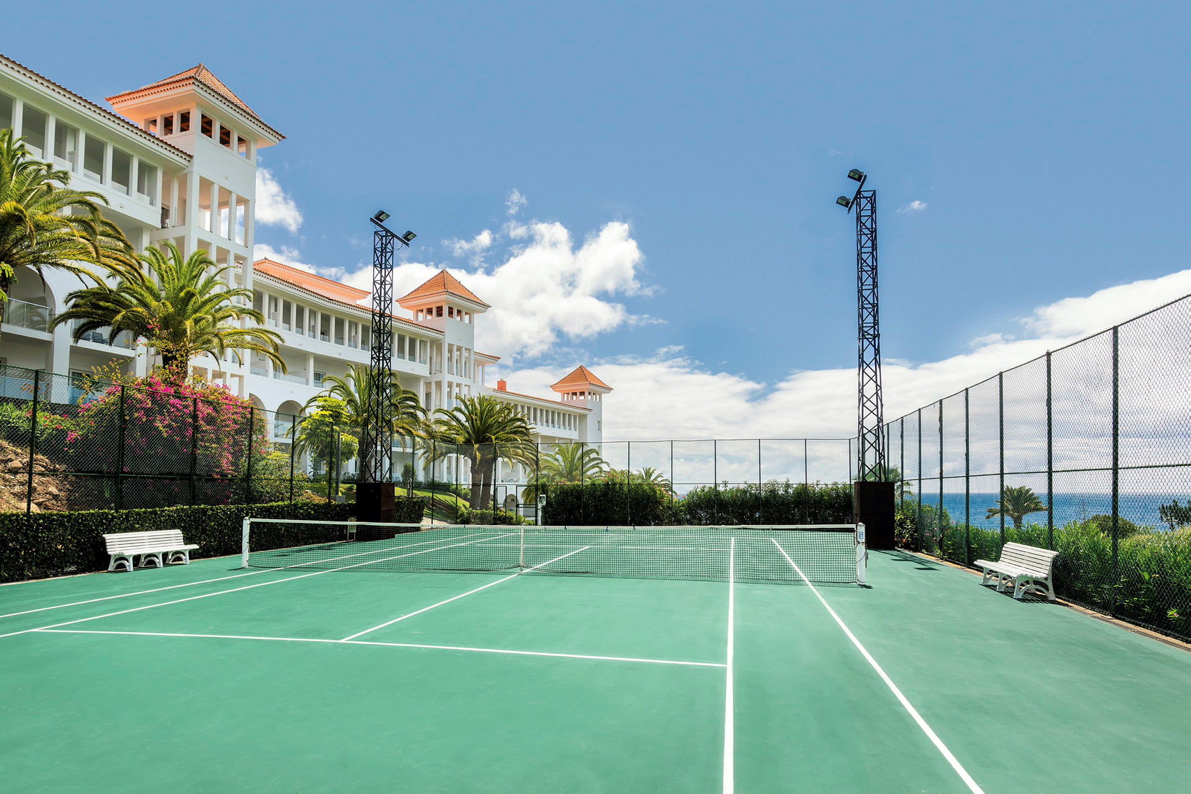 En tennisbana med grön mark och vit linjemarkering omges av stängsel. I bakgrunden syns en vit byggnad med orange tak och palmer samt en blå himmel. (AI-genererad text)
