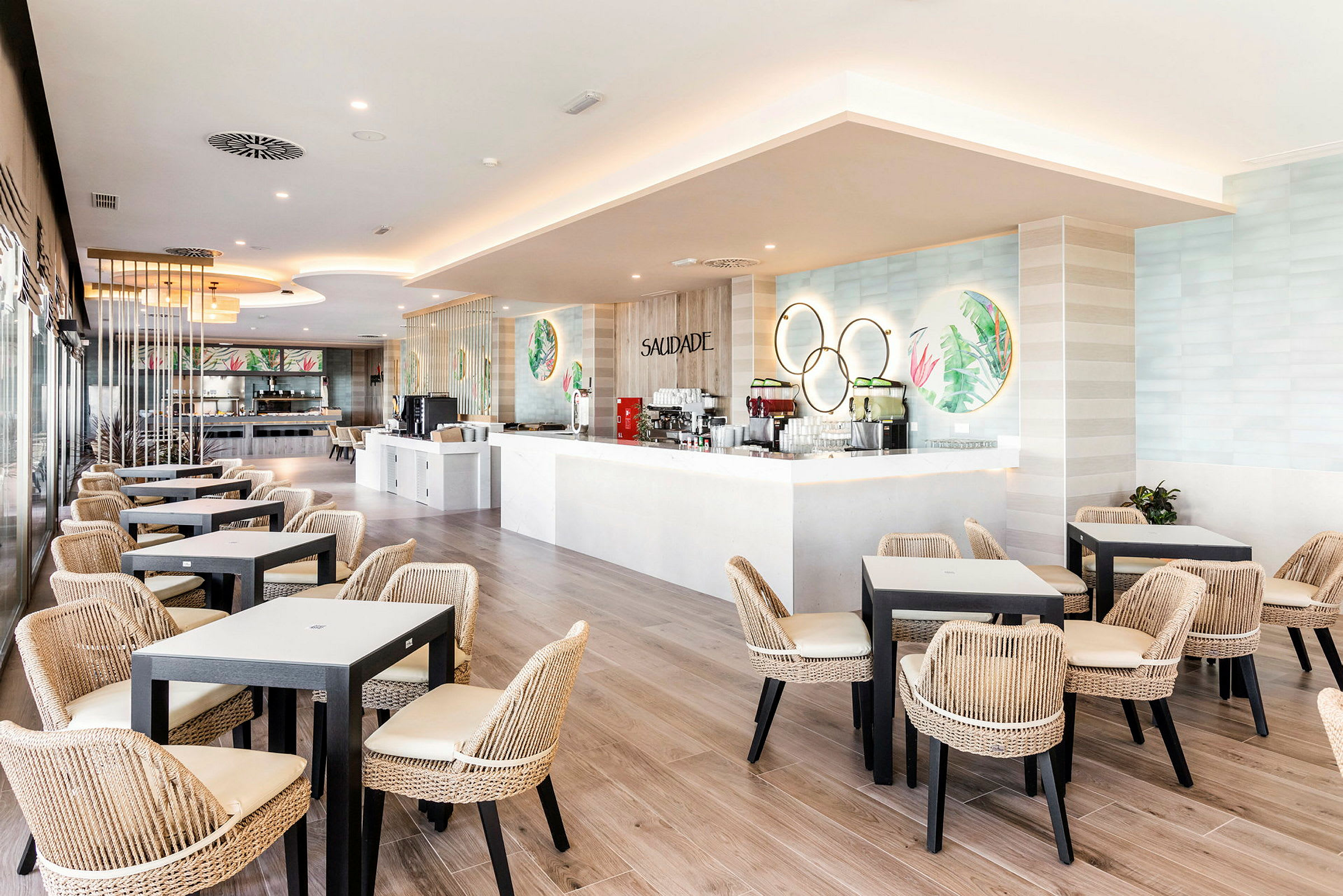 Ljus restaurang med moderna bord och flätade stolar, samt en vit bardisk. (AI-genererad text)
