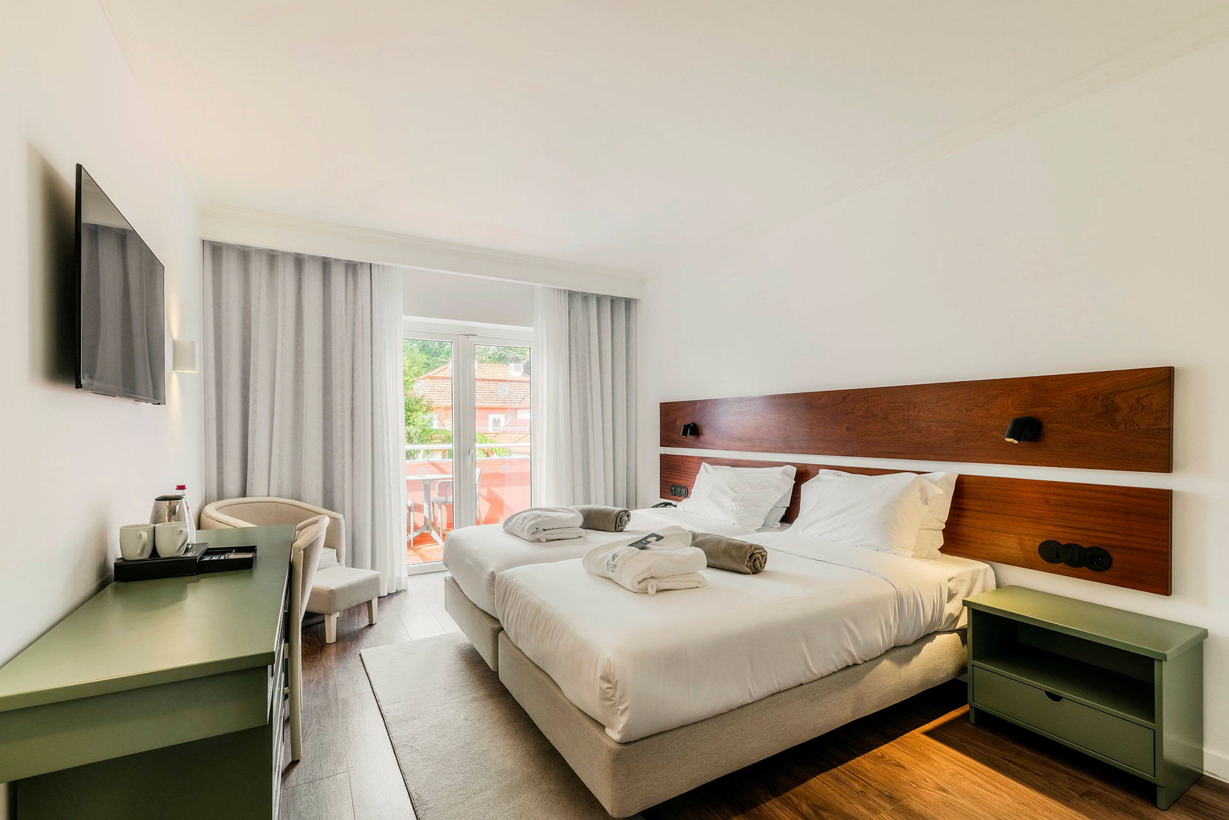 Ett hotellrum med två enkelsängar, vita sängkläder och kuddar. Rummet har en balkong med utsikt och är inrett med ett skrivbord och en TV. (AI-genererad text)