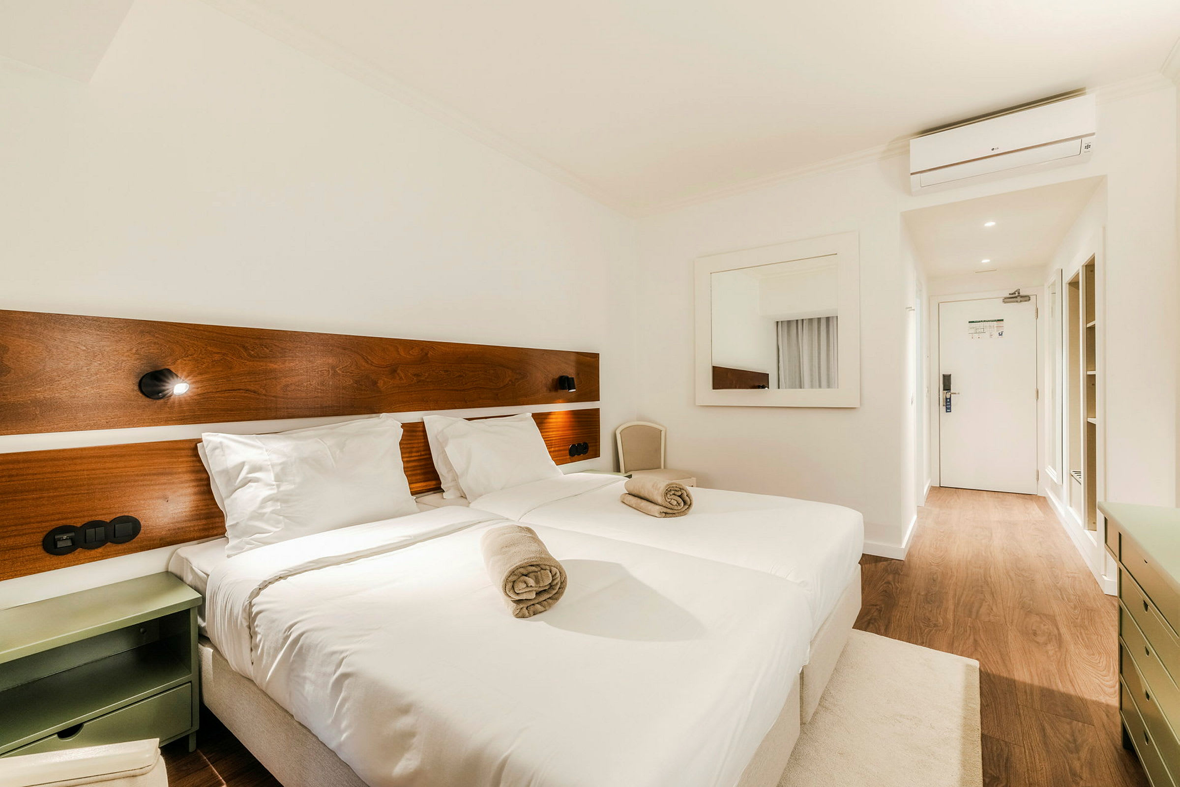 Ett hotellrum med två sängar som har vita lakan och två hoprullade handdukar på. Vägglampor är monterade ovanför sängarna på en träpanel. Ett spegel är uppsatt på väggen och dörren syns i bakgrunden med en träbeloaggolv. (AI-genererad text)