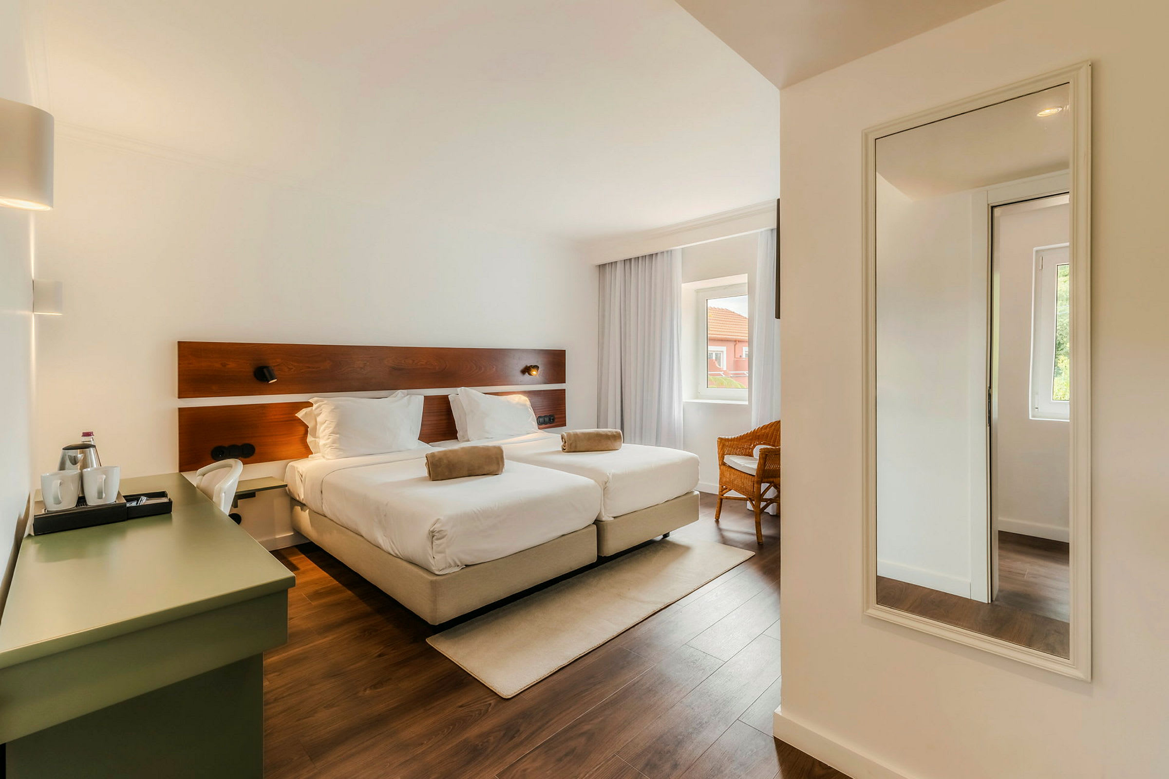 Ett hotellrum med två enkelsängar, en skrivbordshörna och en spegel på väggen. Rummet har ett stort fönster med gardiner. (AI-genererad text)