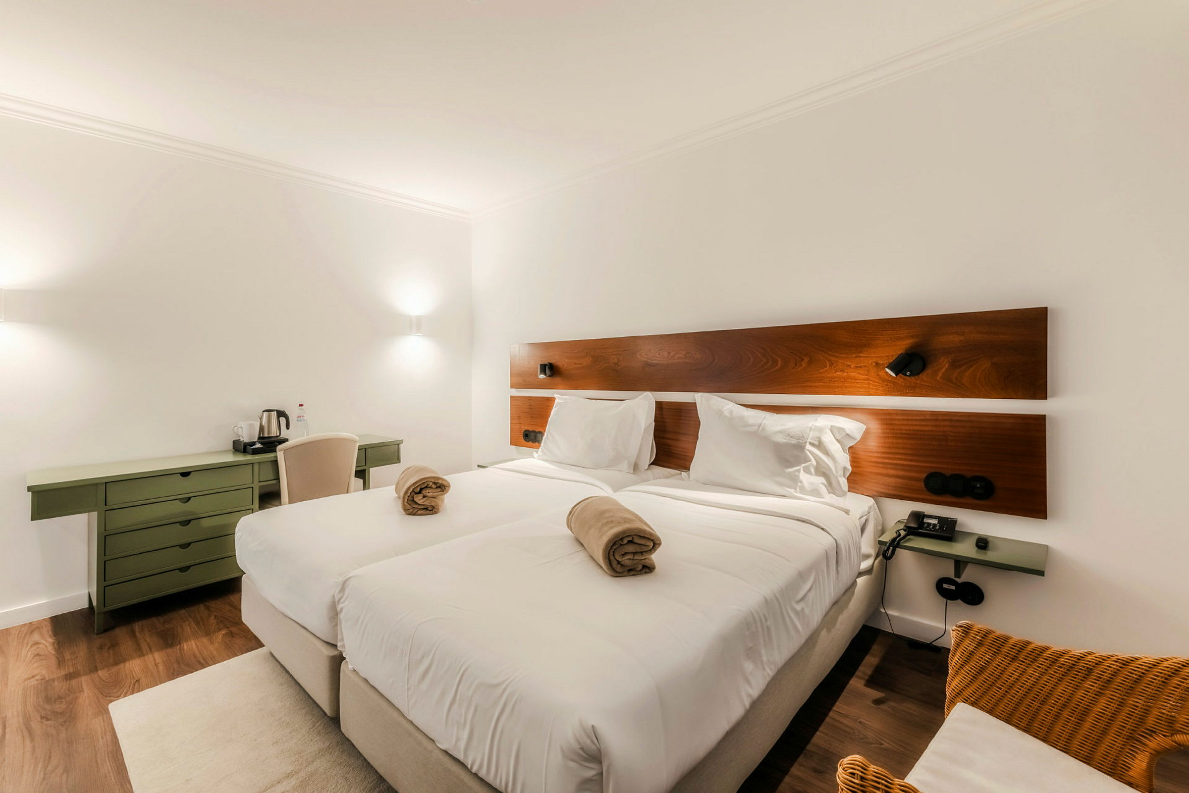 Ett hotellrum med två sammanfogade enkelsängar bäddade med vita sängkläder. Över sängarna finns en träpanel på väggen och rummet är inrett med ett grönt skrivbord och en stol. (AI-genererad text)