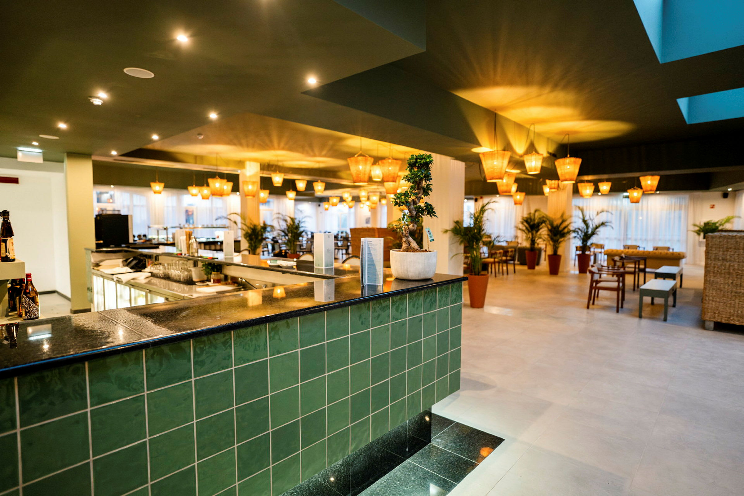 En interiör av en modern restaurang med grön kakel, en bardisk och flera bord med stolar samt växter. (AI-genererad text)