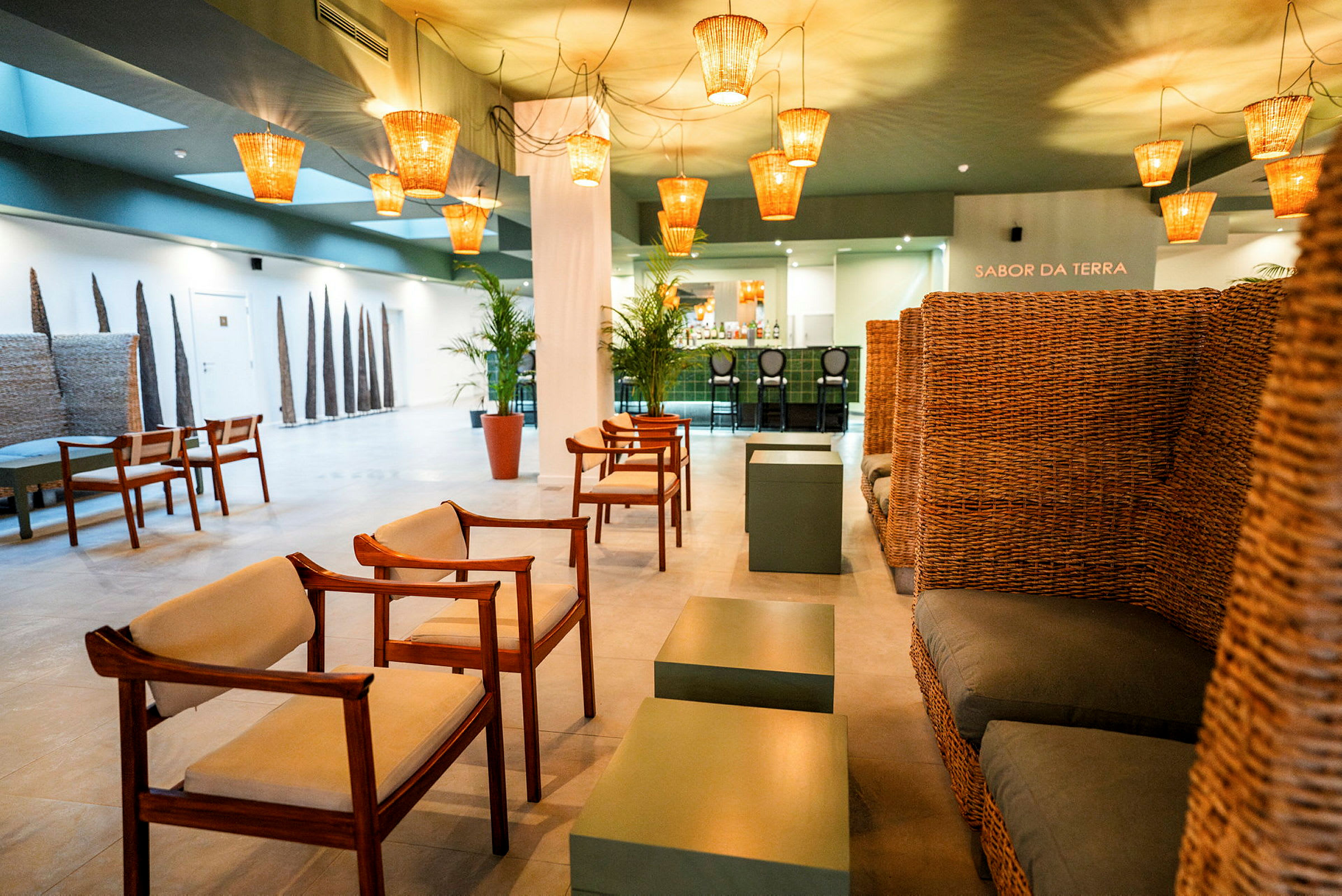 En hotellets lounge med trästolar, soffor flätade i rotting, gröna bord och varm belysning från takhängande lampor. (AI-genererad text)