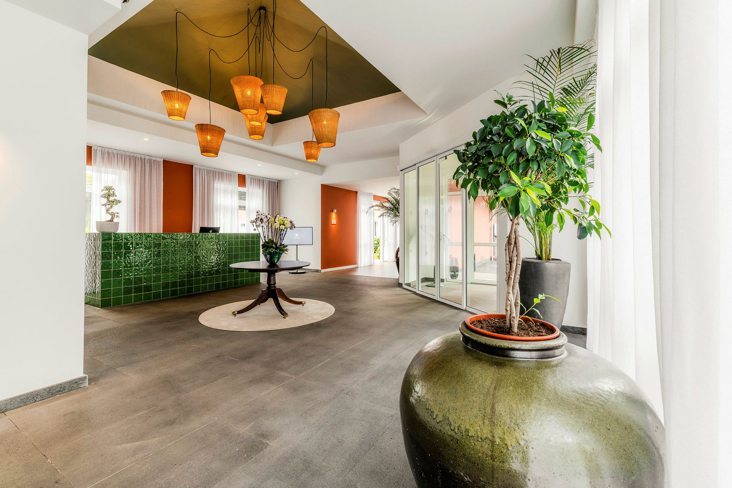 En ljus och modern hotellobby med gröna keramiska detaljväggar, en rund bord med blommor, samt stora krukväxter i förgrunden. (AI-genererad text)