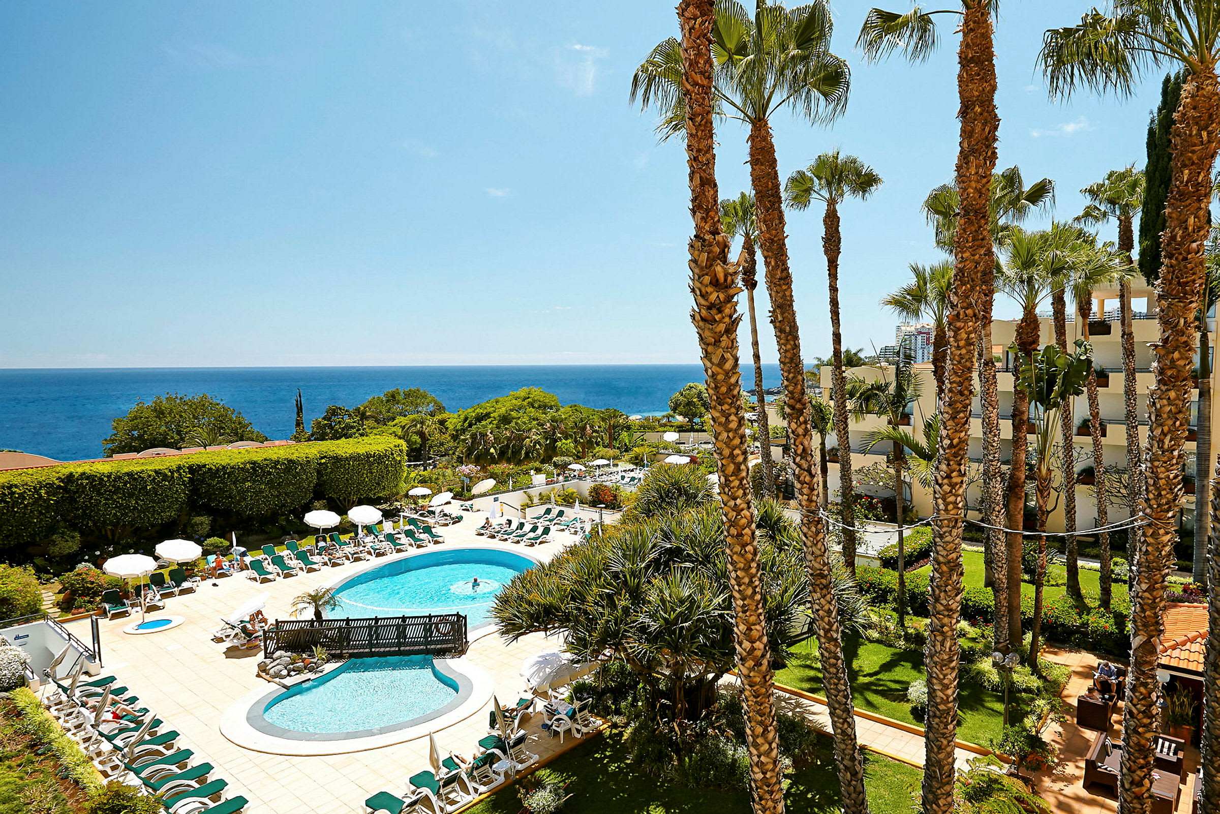 En hotellpool med solstolar och parasoller är omgiven av grönska med havet i bakgrunden. (AI-genererad text)