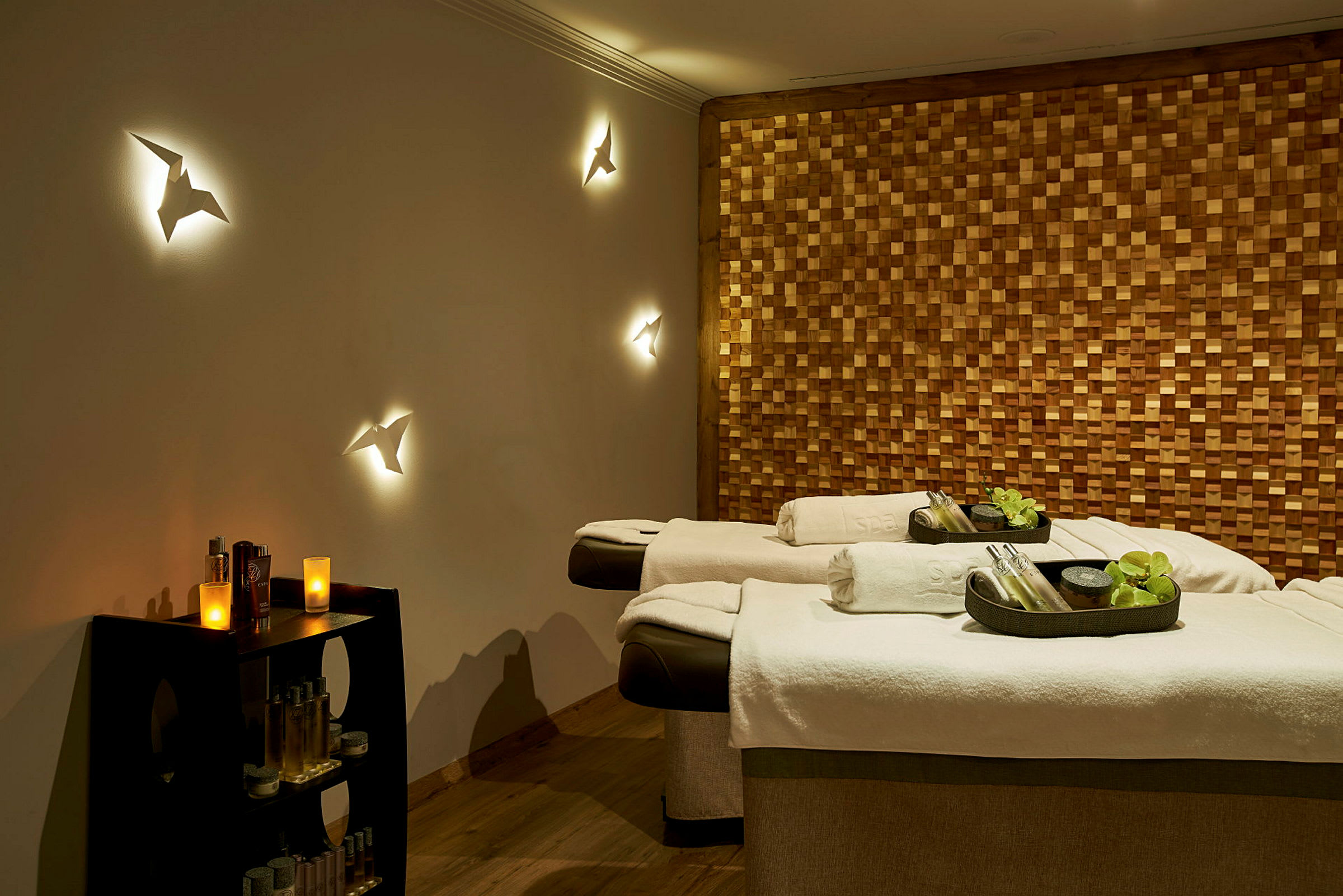 Ett spa-rum med massagebänkar förberedda med vita handdukar och dekorationer. En vägg är beklädd med träpaneler, medan ljus och prydnader skapar en avslappnande atmosfär. (AI-genererad text)