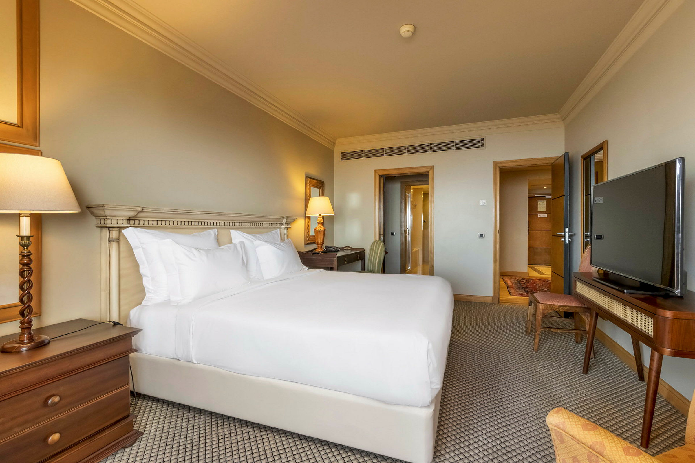 Ett hotellrum med en dubbelsäng täckt av vitt sänglinne, en nattduksbordslampa och en TV på ett sidobord. (AI-genererad text)