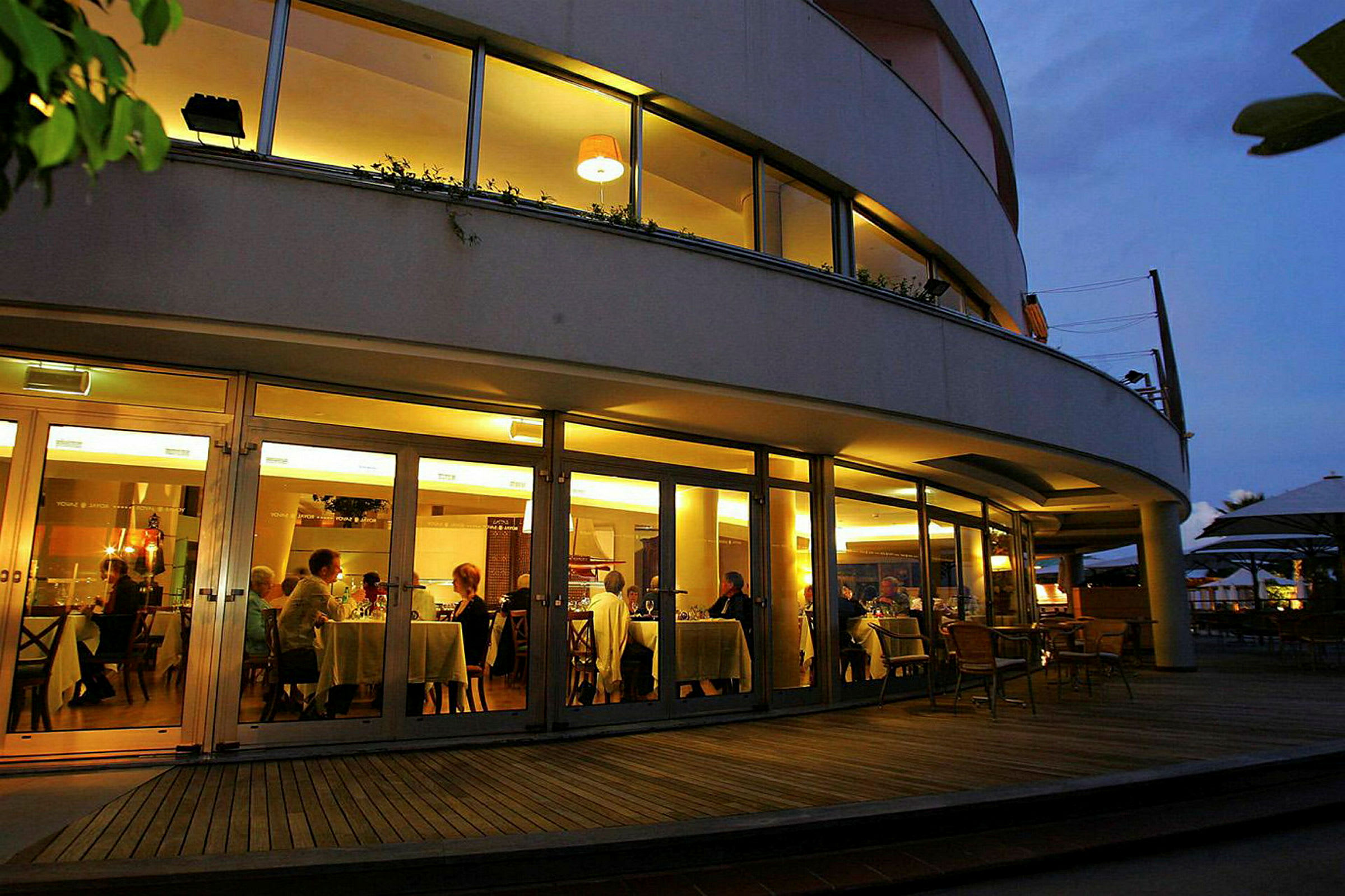 En restaurang med stora glasfönster genom vilka personer vid borden kan ses samlade till middag. (AI-genererad text)