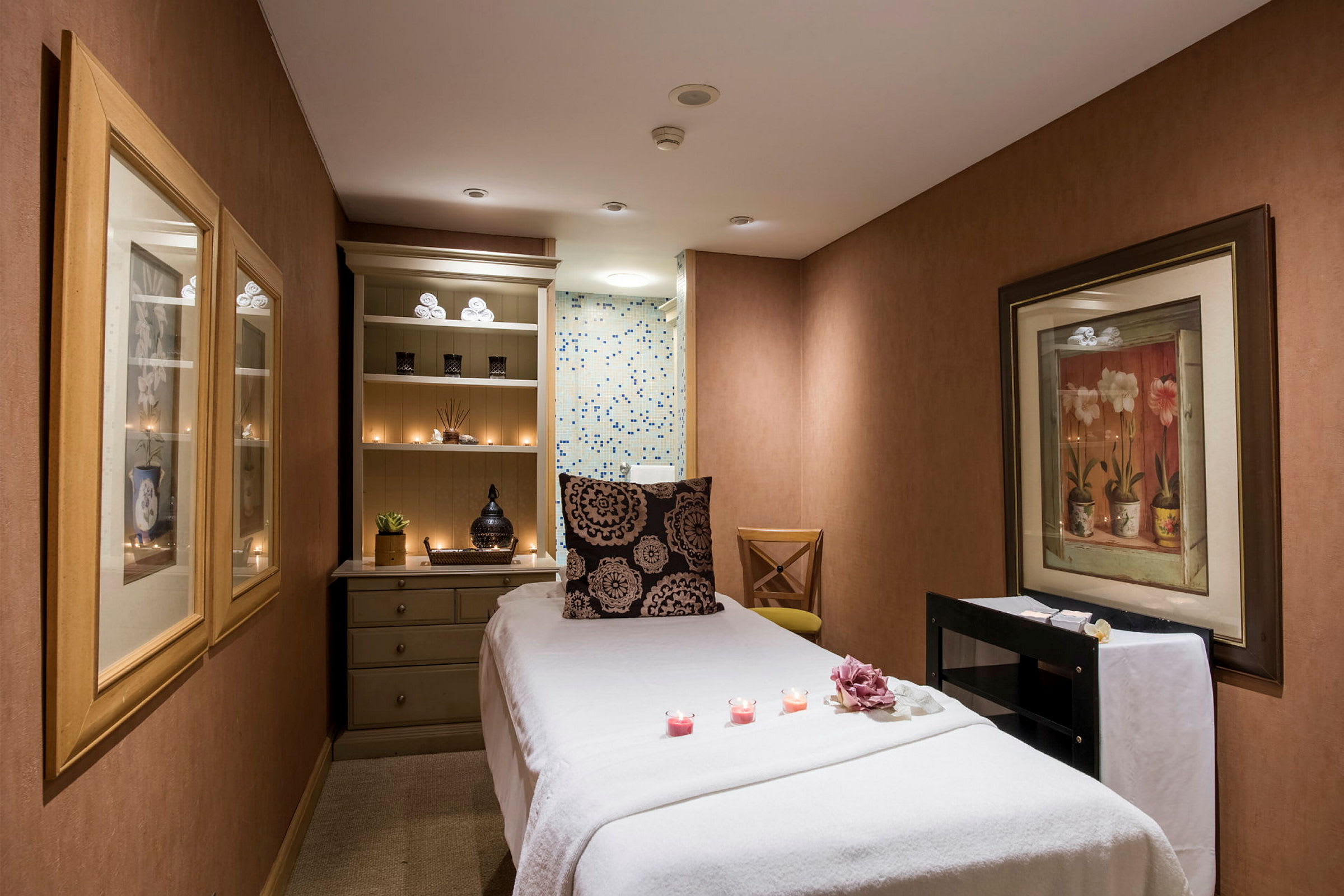 Ett spa-rum med en massagebänk, omgiven av dekorativa element såsom ljus och konstverk på väggarna. (AI-genererad text)