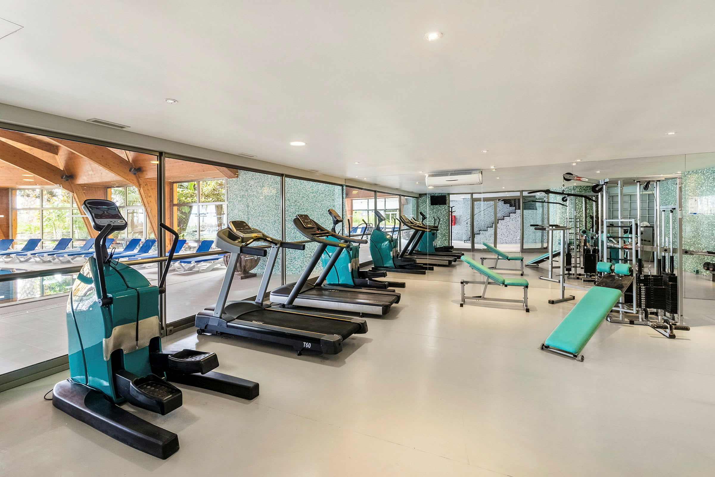 Gym med träningsutrustning som crosstrainers, löpband och styrketräningsmaskiner, bredvid stora fönster med utsikt över en pool. (AI-genererad text)