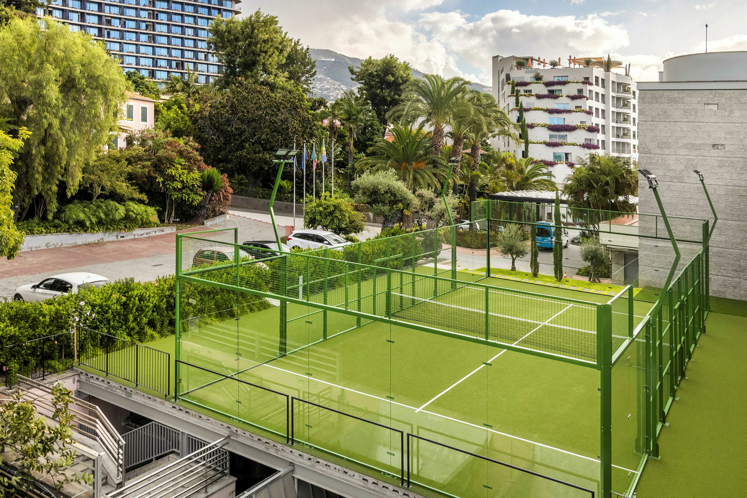 En grön padelbana med glasväggar omgiven av träd och byggnader. (AI-genererad text)