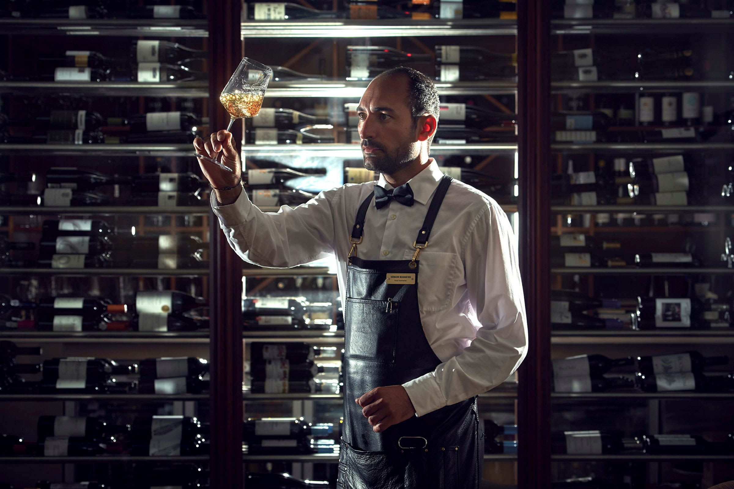 Bartender undersöker innehållet i ett vinglas framför en vinförvaringshylla. (AI-genererad text)