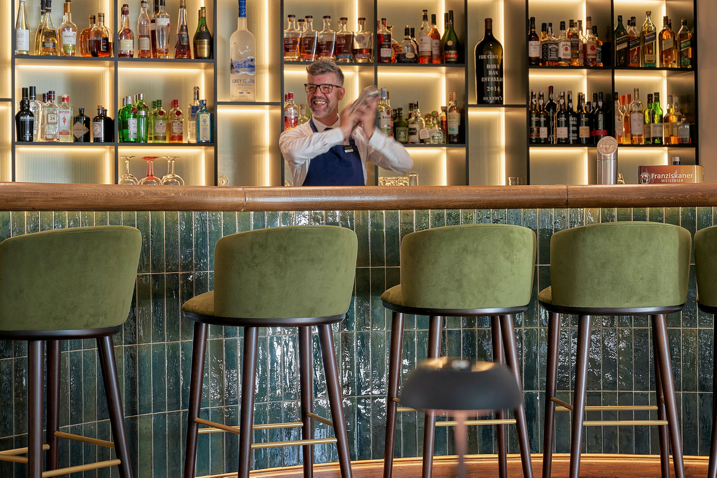 En bar med gröna barstolar och en hylla med flaskor i bakgrunden. (AI-genererad text)