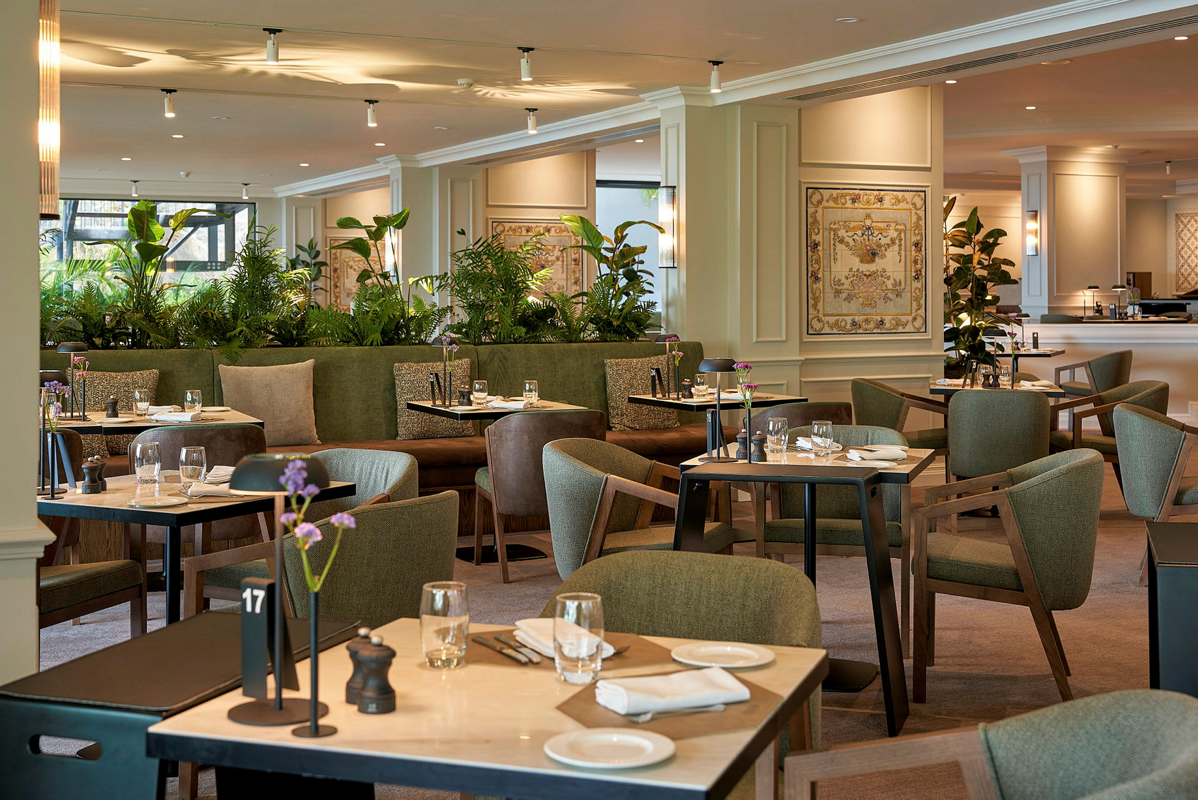 Ett elegant restaurangområde med grön inredning, bord dukade för måltider och dekorativa växtarrangemang. (AI-genererad text)